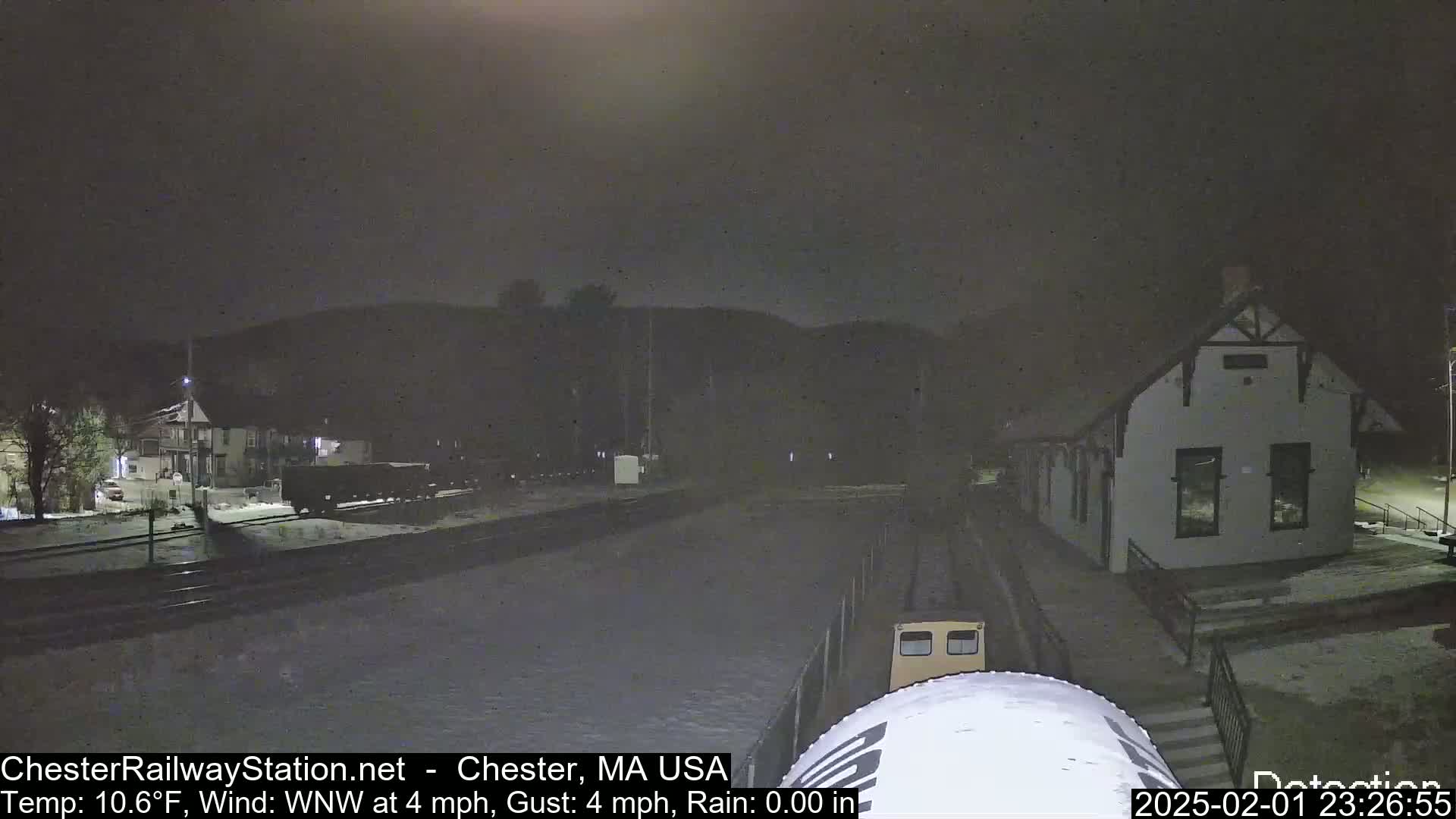 A nighttime view of a train station and railway tracks covered in snow, with a train partially visible, under a dark sky.