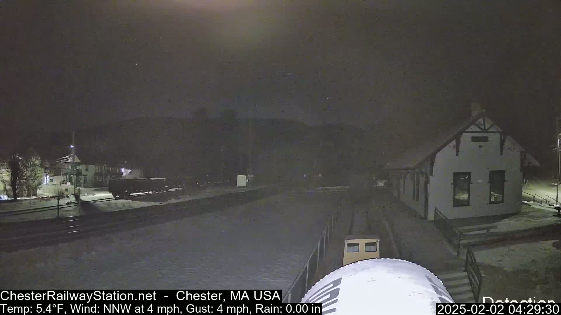 A nighttime view of a train station and railway tracks in snowy conditions, with a train car partially visible in the foreground.