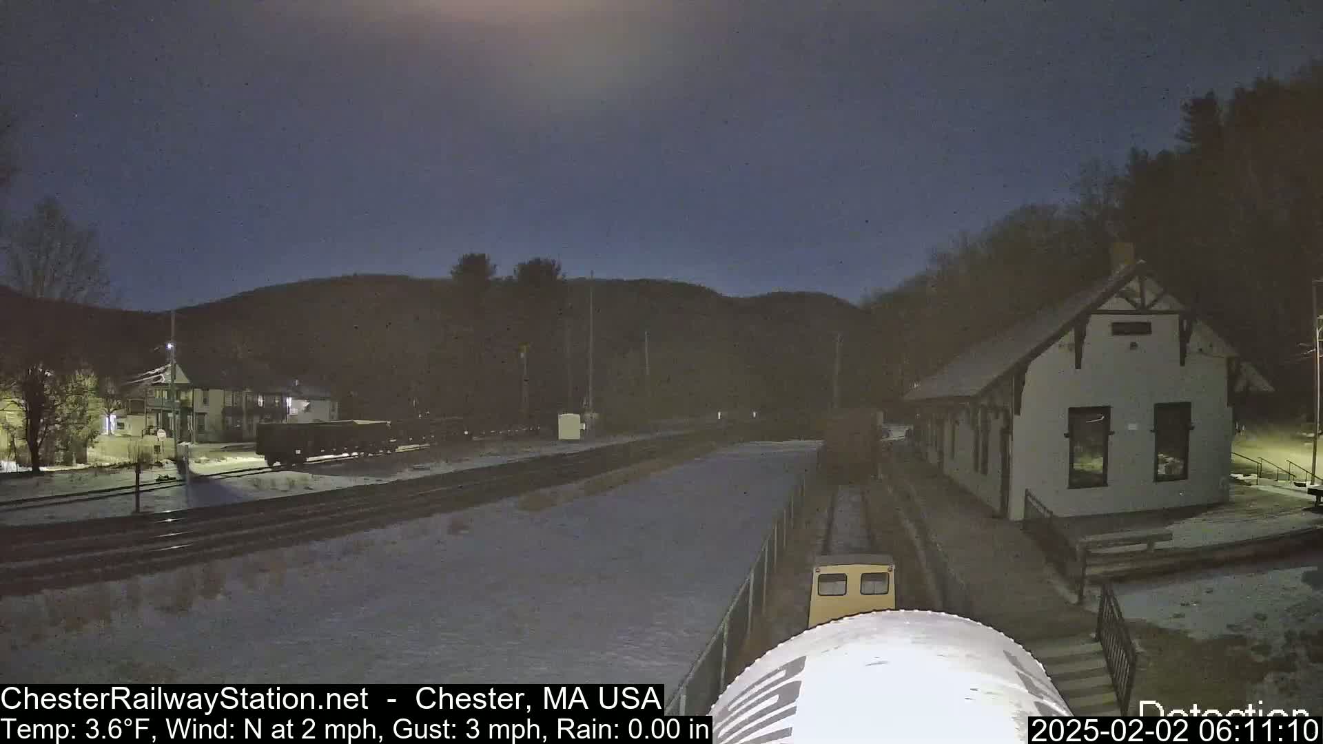 A nighttime view of a train station and rail yard under a dark, clear sky, with snow on the ground.