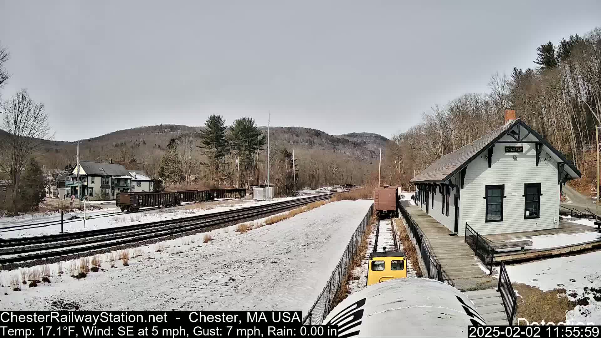 A snow-covered train station and railway tracks are visible, with a small train approaching the station and hills in the background under a cloudy sky.