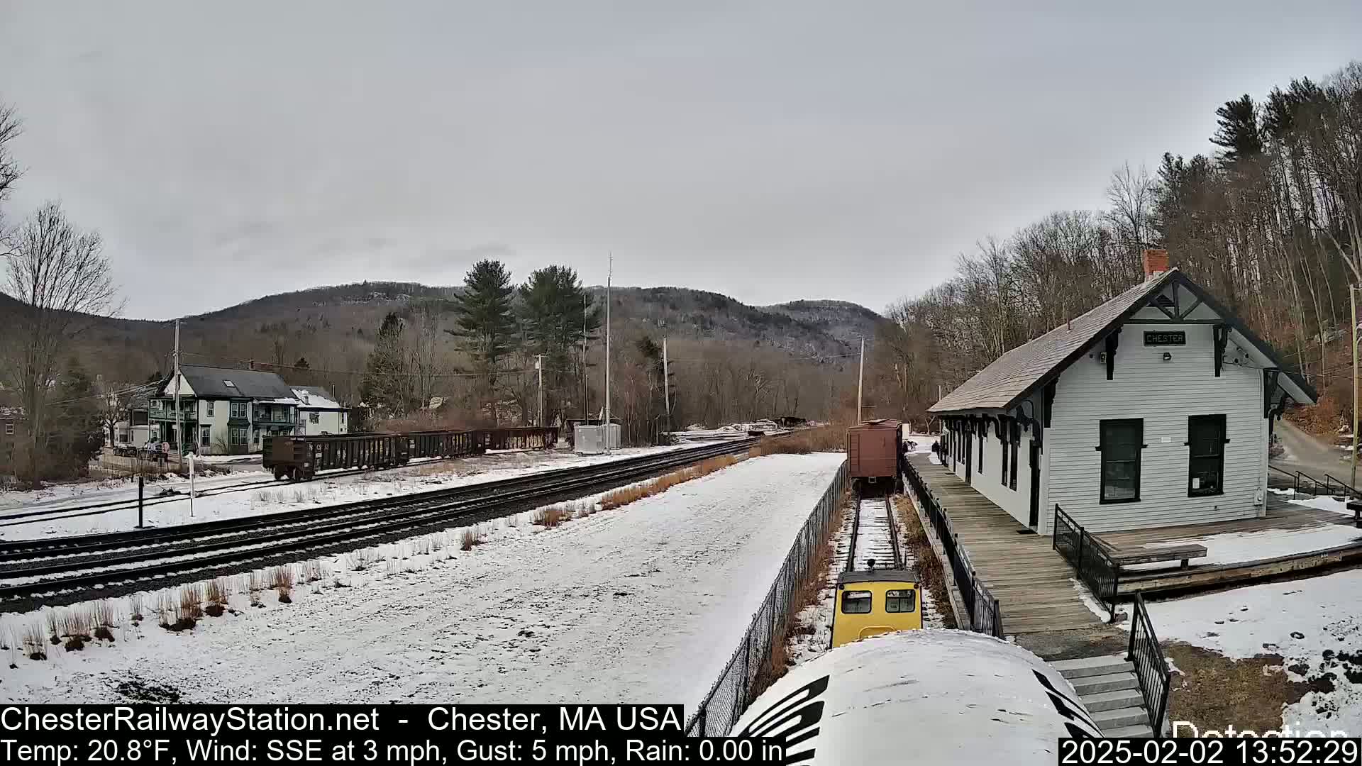 A snow-covered train station and railway tracks are seen under an overcast sky, with a small yellow train car approaching the station and hills in the background.