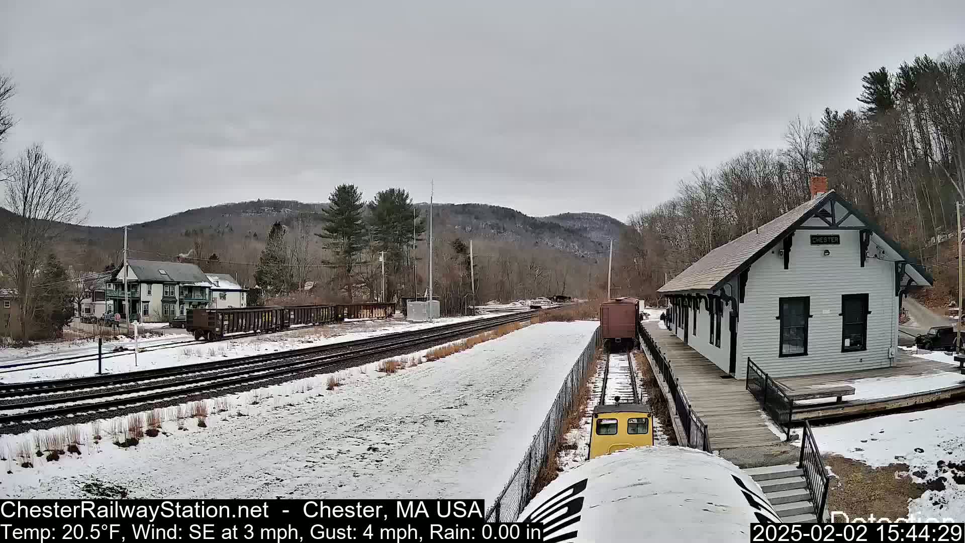 A snow-covered train station and tracks sit against a backdrop of bare trees and hills under an overcast sky.