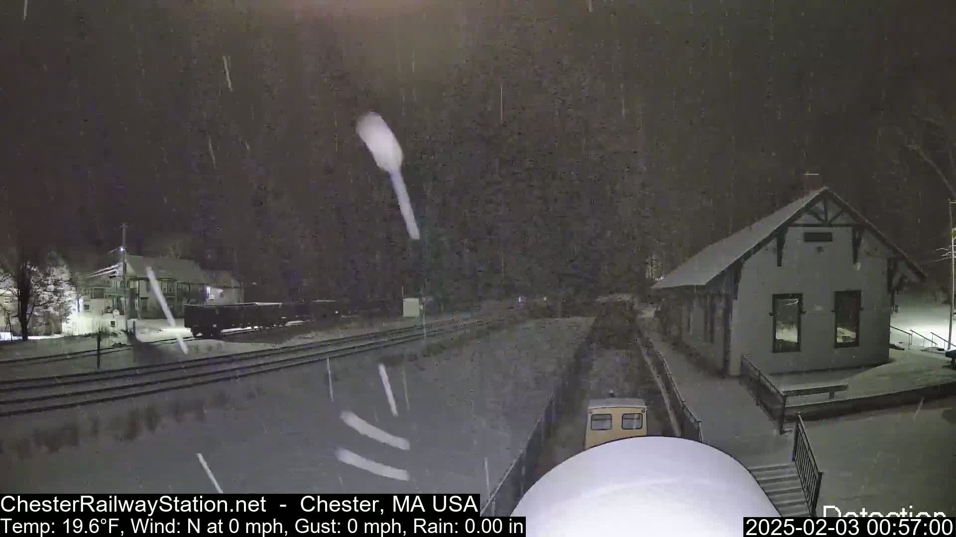 Snow falls at night on a train station and railway tracks.