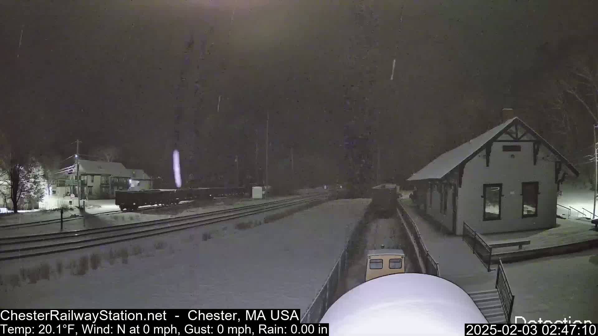 A snow-covered train station and railway yard are visible under a nighttime snowfall.