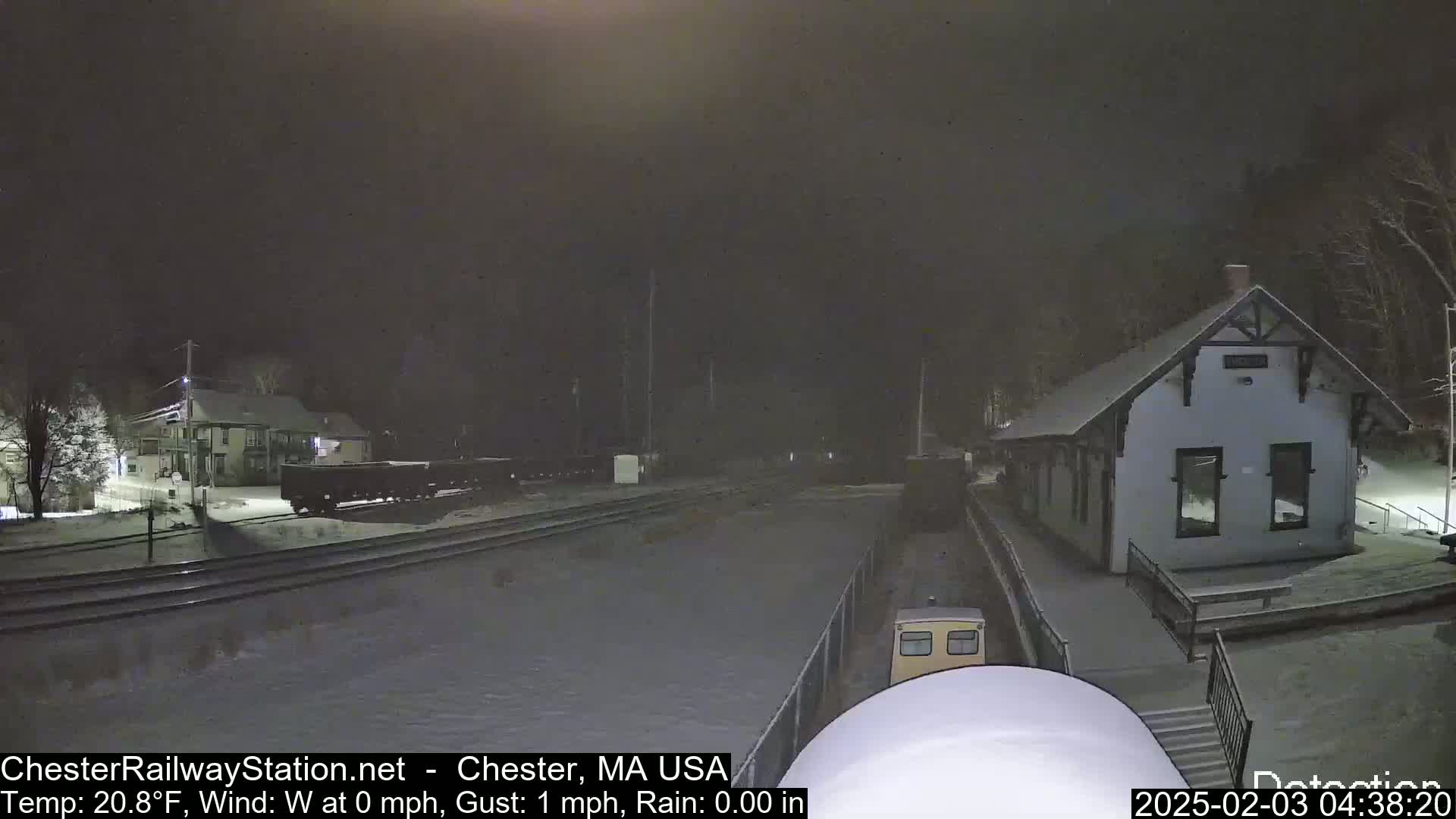 A snow-covered train station and rail yard are visible at night under light snowfall.