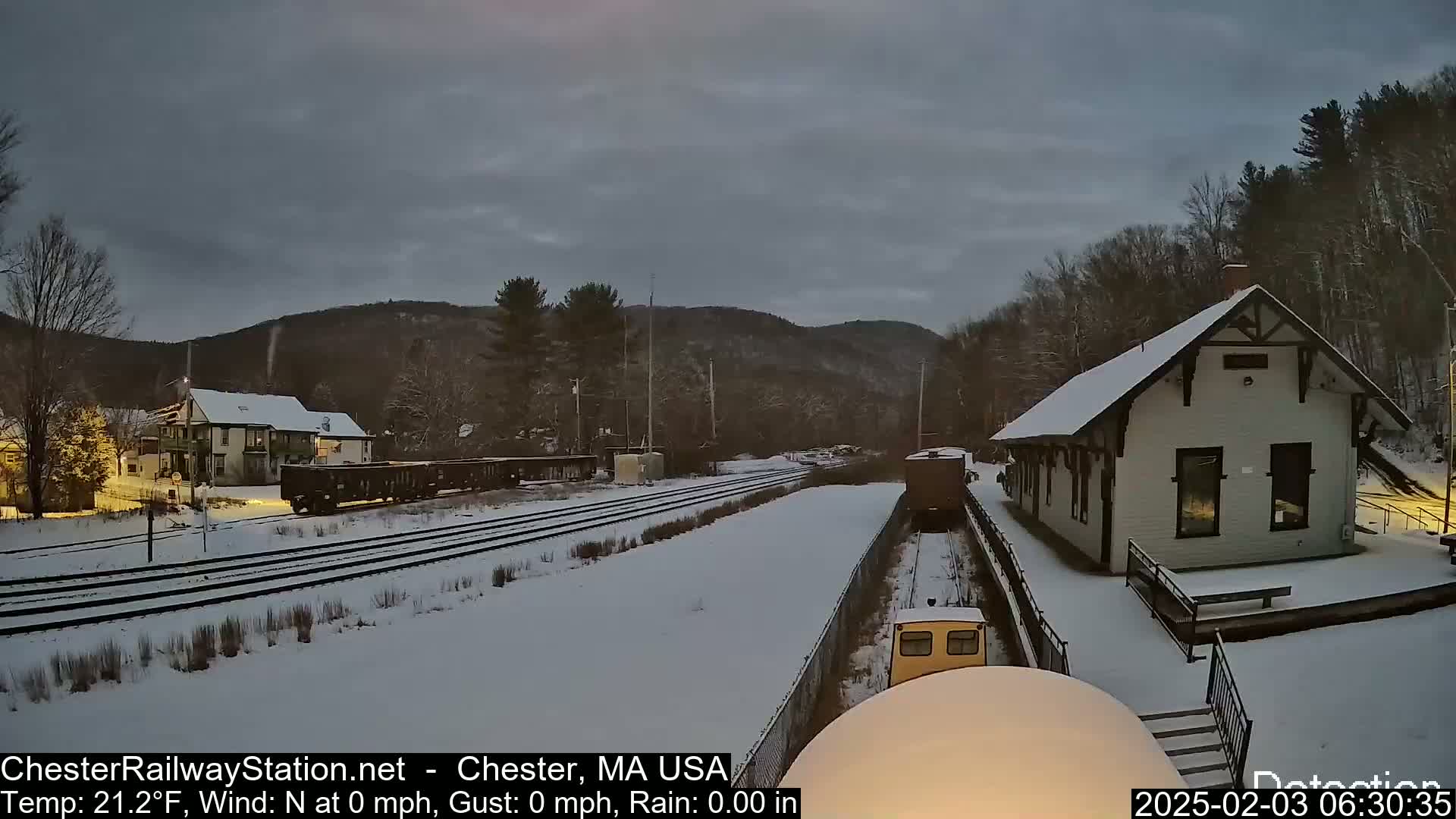 A snow-covered train station and railway tracks are visible under a cloudy sky, with a small train approaching the station.