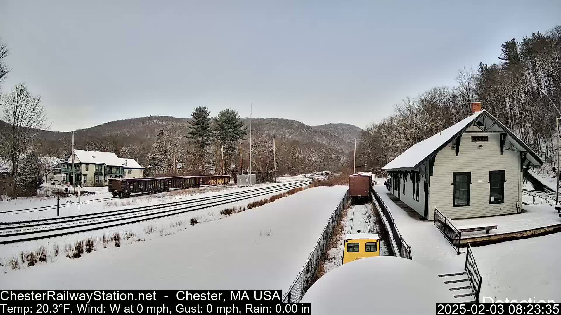 A snow-covered train station and railway yard are nestled in a hilly, wooded landscape under a cloudy sky.