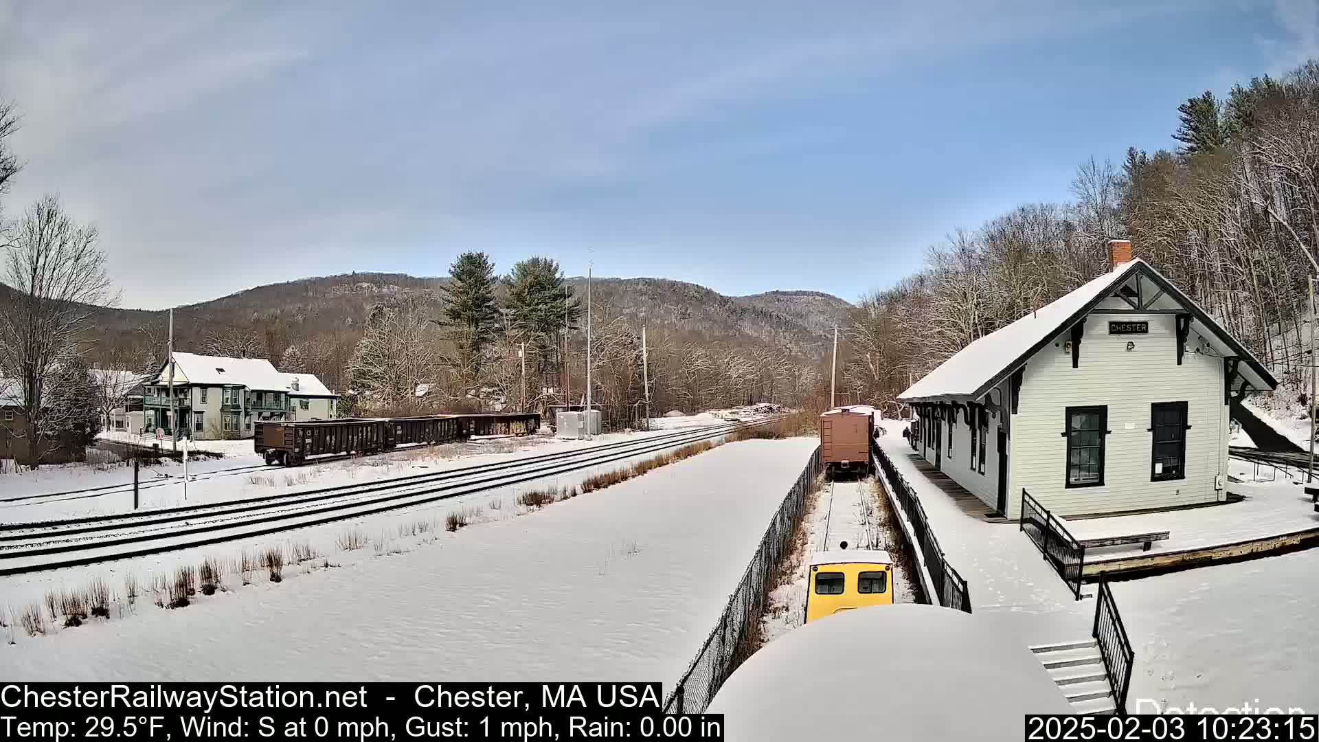 A snow-covered train station and rail yard are seen on a partly sunny day, with a small yellow train car in the foreground and freight cars further down the tracks.