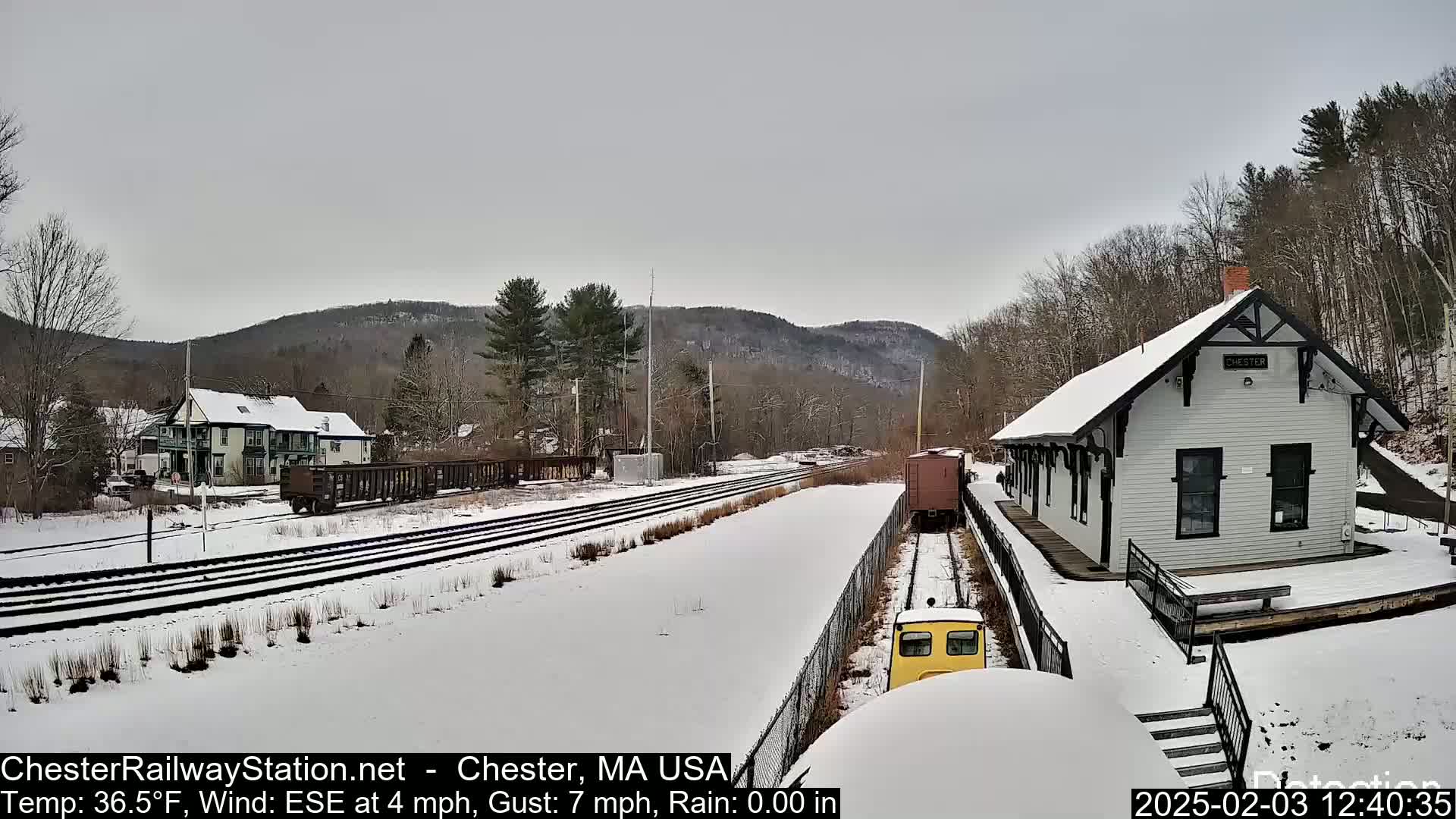 A snow-covered train station and railway tracks are seen, with a small yellow train car partially visible in the foreground, under a cloudy sky.