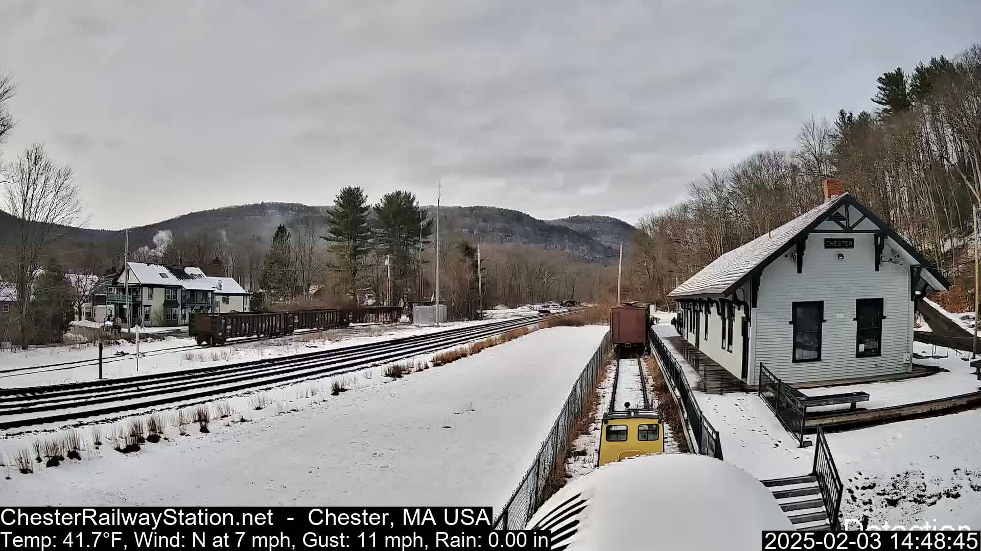 A snow-covered train station and railroad tracks are visible, with a small yellow train car and a freight car in the foreground, under an overcast sky.