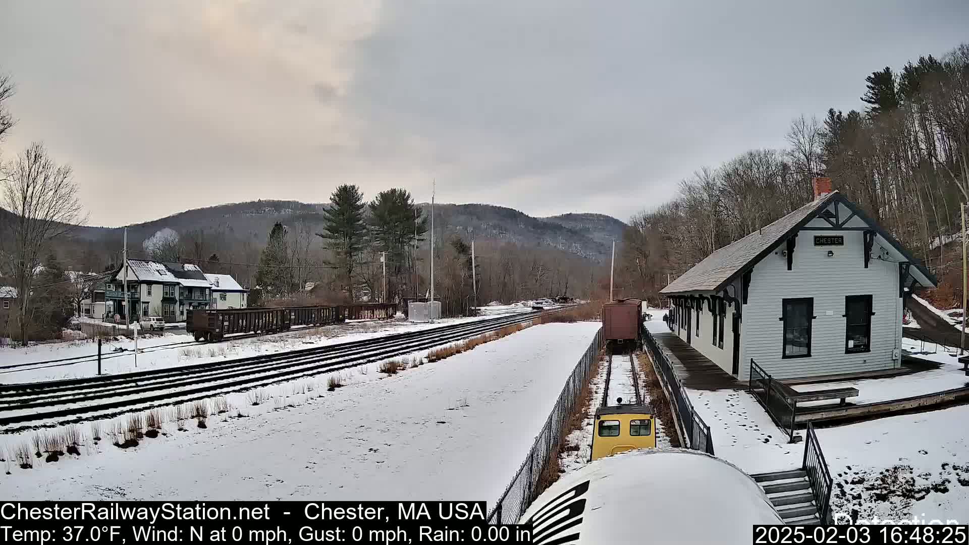 A snow-covered train station and rail yard are visible, with a small train approaching the station under a cloudy sky.