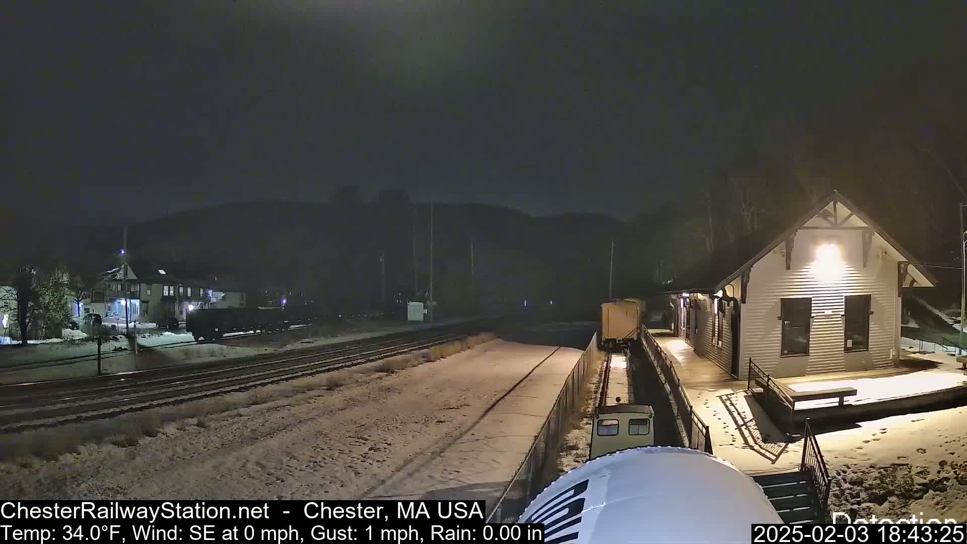 A snow-covered train station at night with a train car nearby and a dark hill in the background under a clear sky.