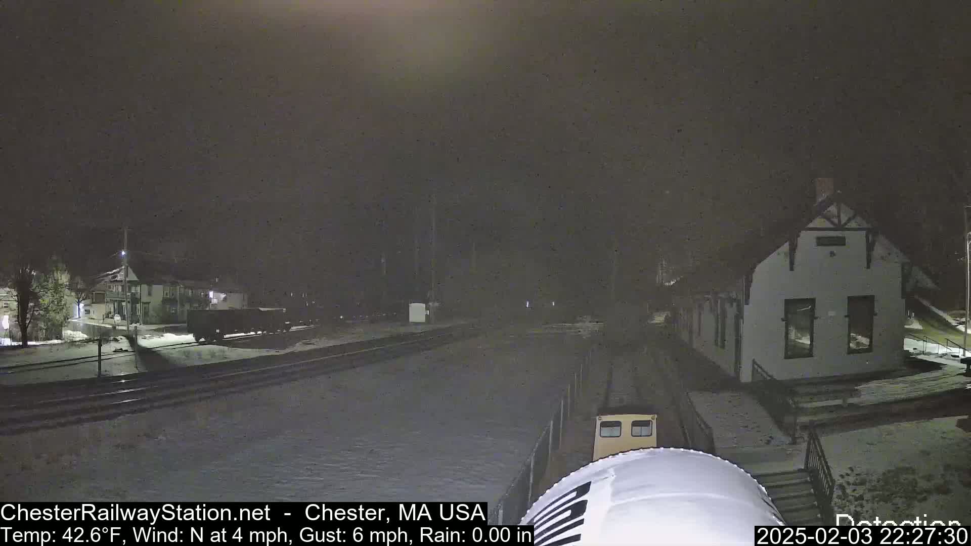 A nighttime view of a train station and railway tracks lightly covered in snow, with a train car partially visible in the foreground under a dark sky.