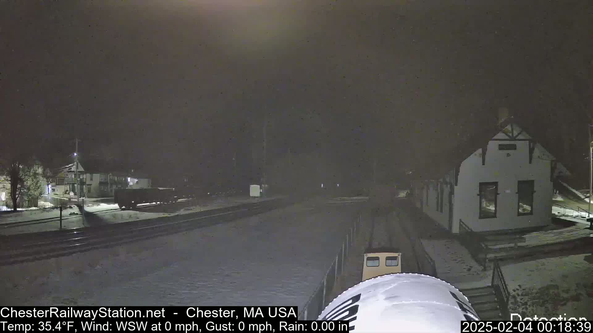 A nighttime view of a snow-covered train station and railroad tracks, with a train partially visible in the foreground.