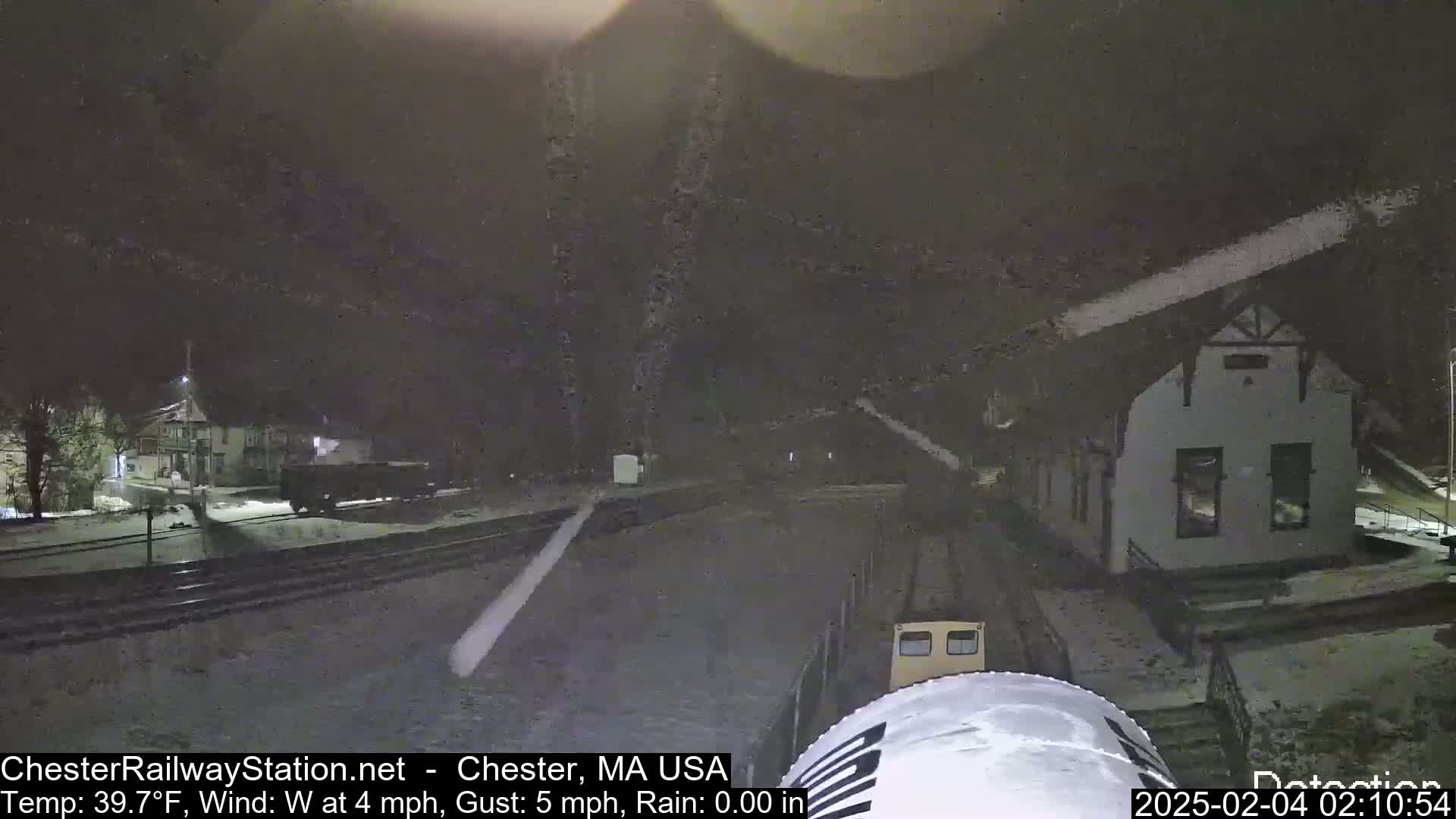 A nighttime view of a train in a snow flurry approaching a small building next to train tracks.