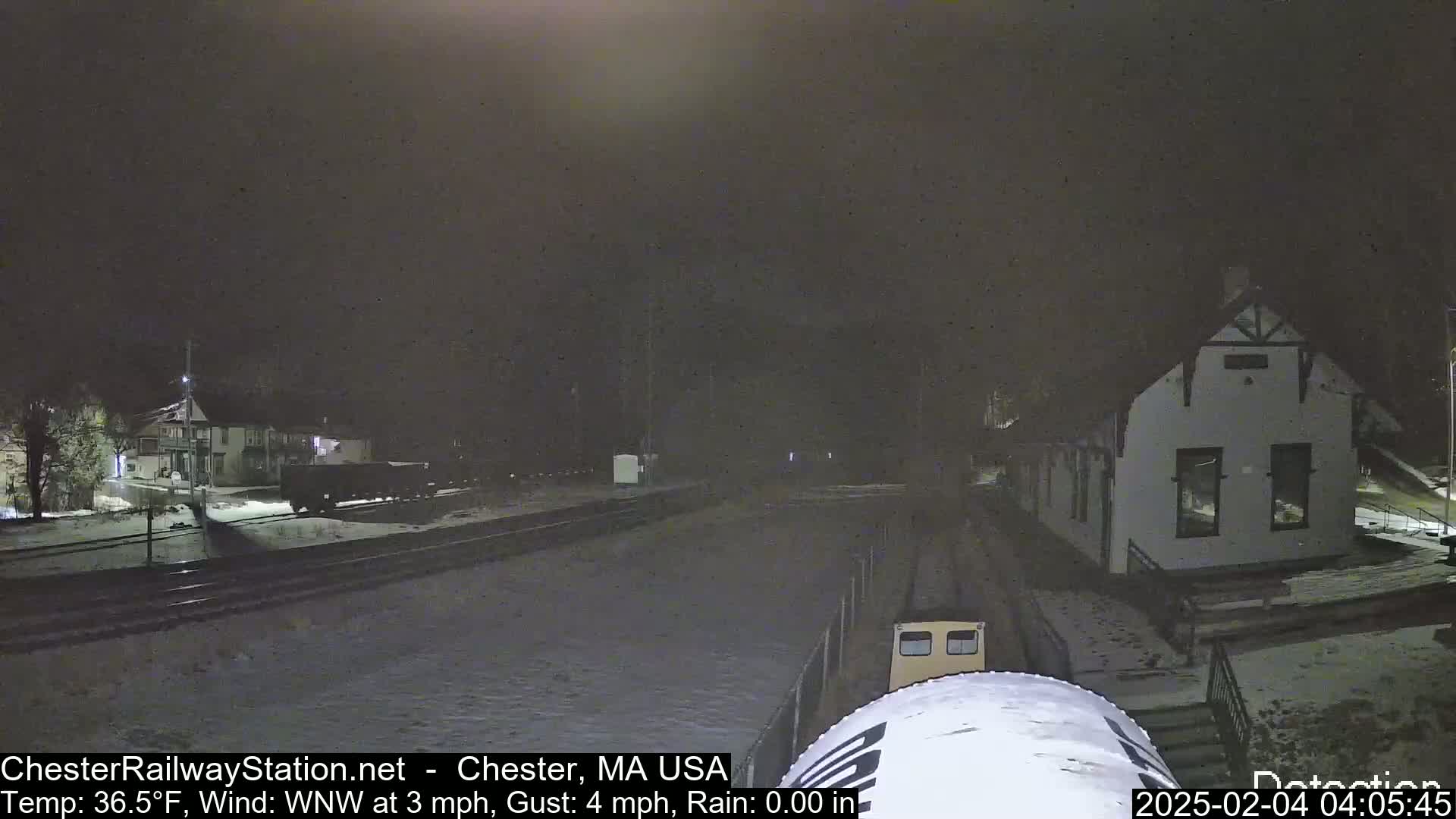 A nighttime view of a snow-covered train station and tracks, with a train car partially visible in the foreground, under a dark sky with a temperature of 36.5°F and a WNW wind.