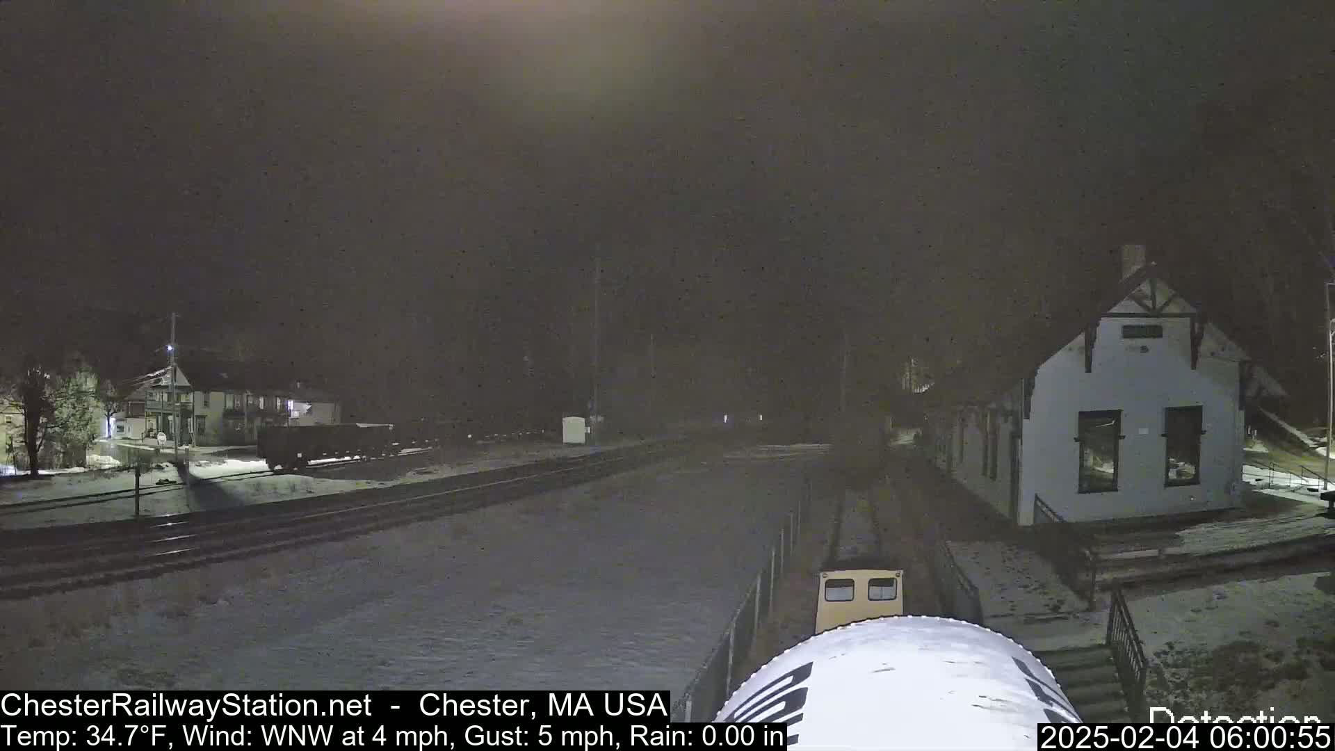 A nighttime scene of a snow-covered train station and railway tracks under a dark sky with light rain.