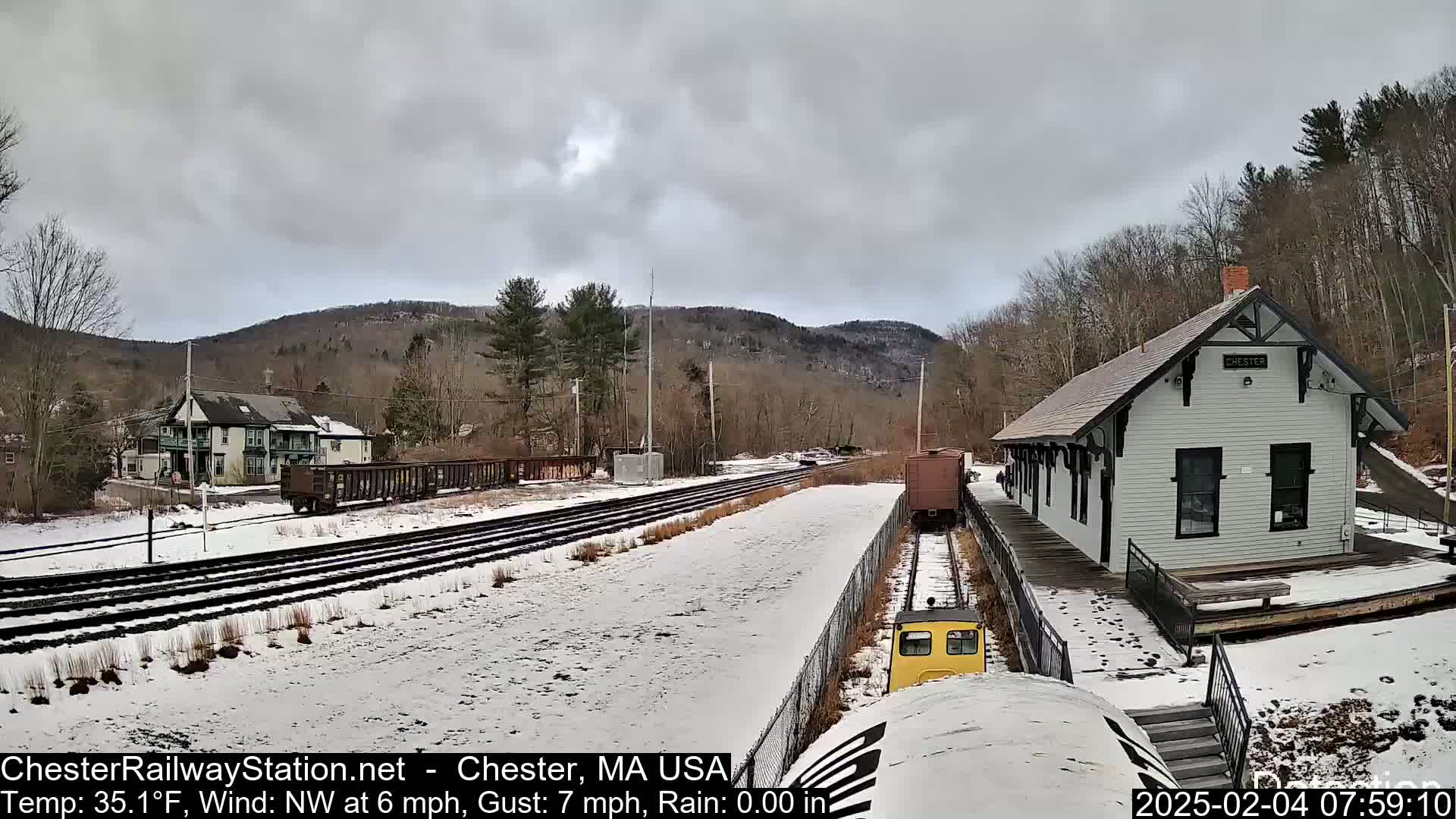 A snow-covered train yard with a small train approaching a white train station situated in a wooded area under a cloudy sky.