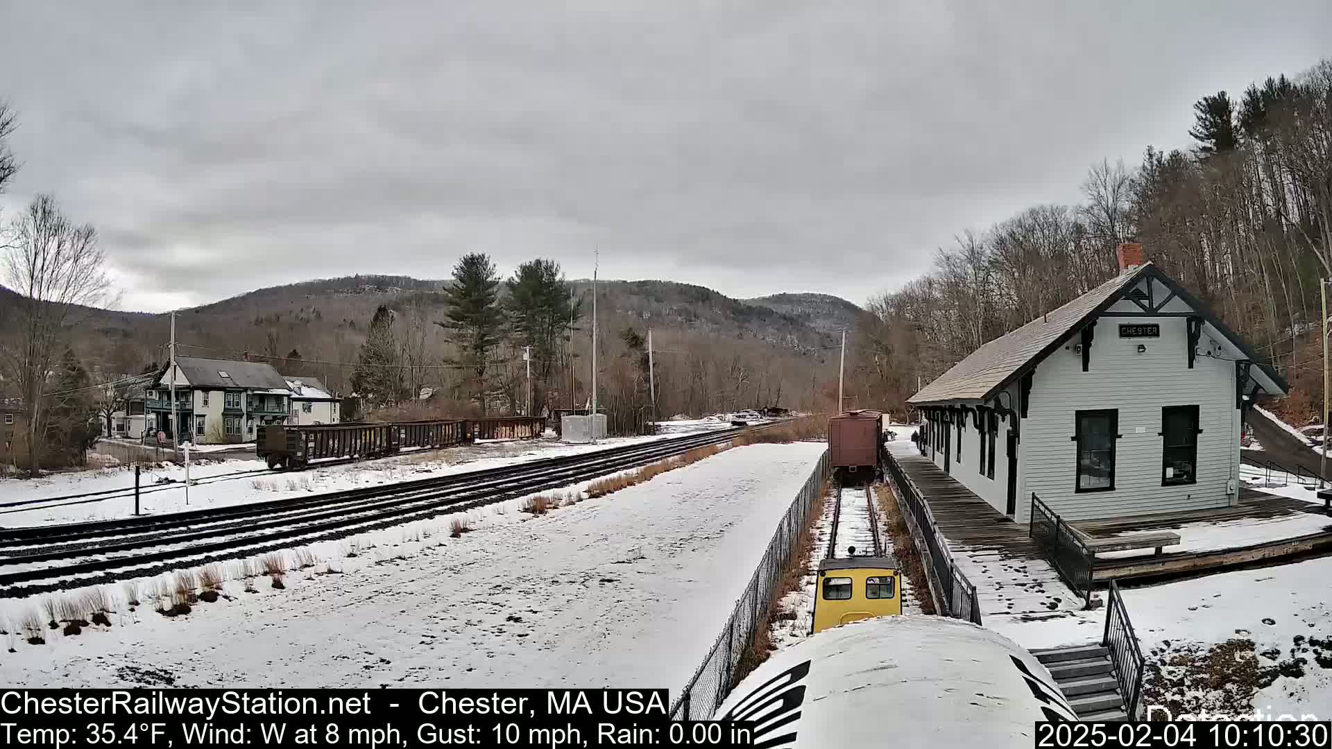 A snow-covered train yard with a small train approaching a white train station building under an overcast sky.