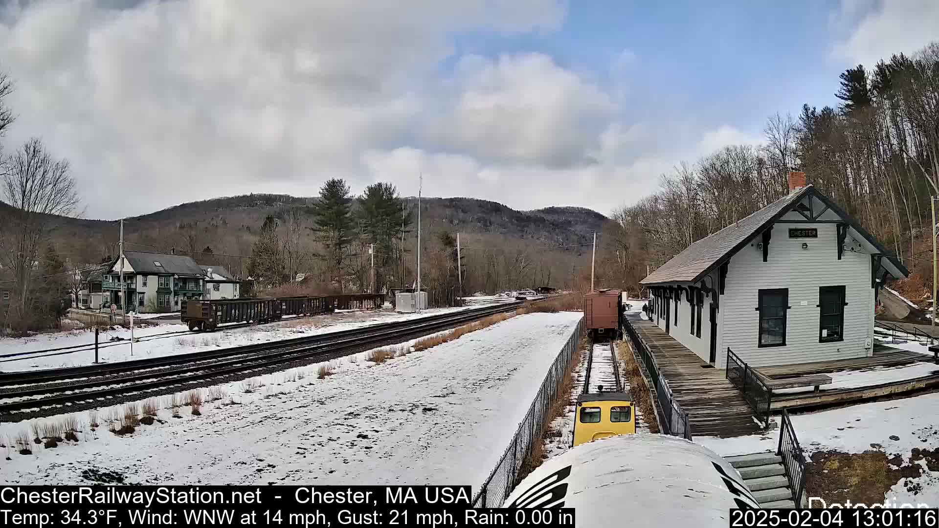 A snow-covered train station and railway yard are visible, with a small yellow train car and several freight cars present under partly cloudy skies.