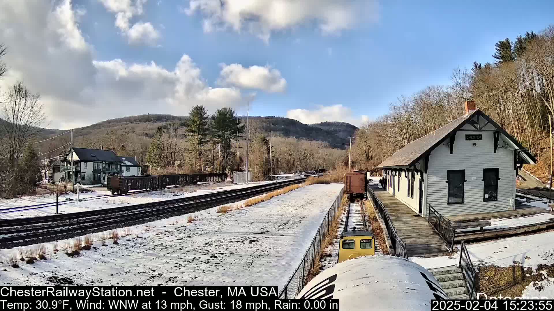 A snow-covered train station and tracks are seen under a partly cloudy sky, with a small train approaching.