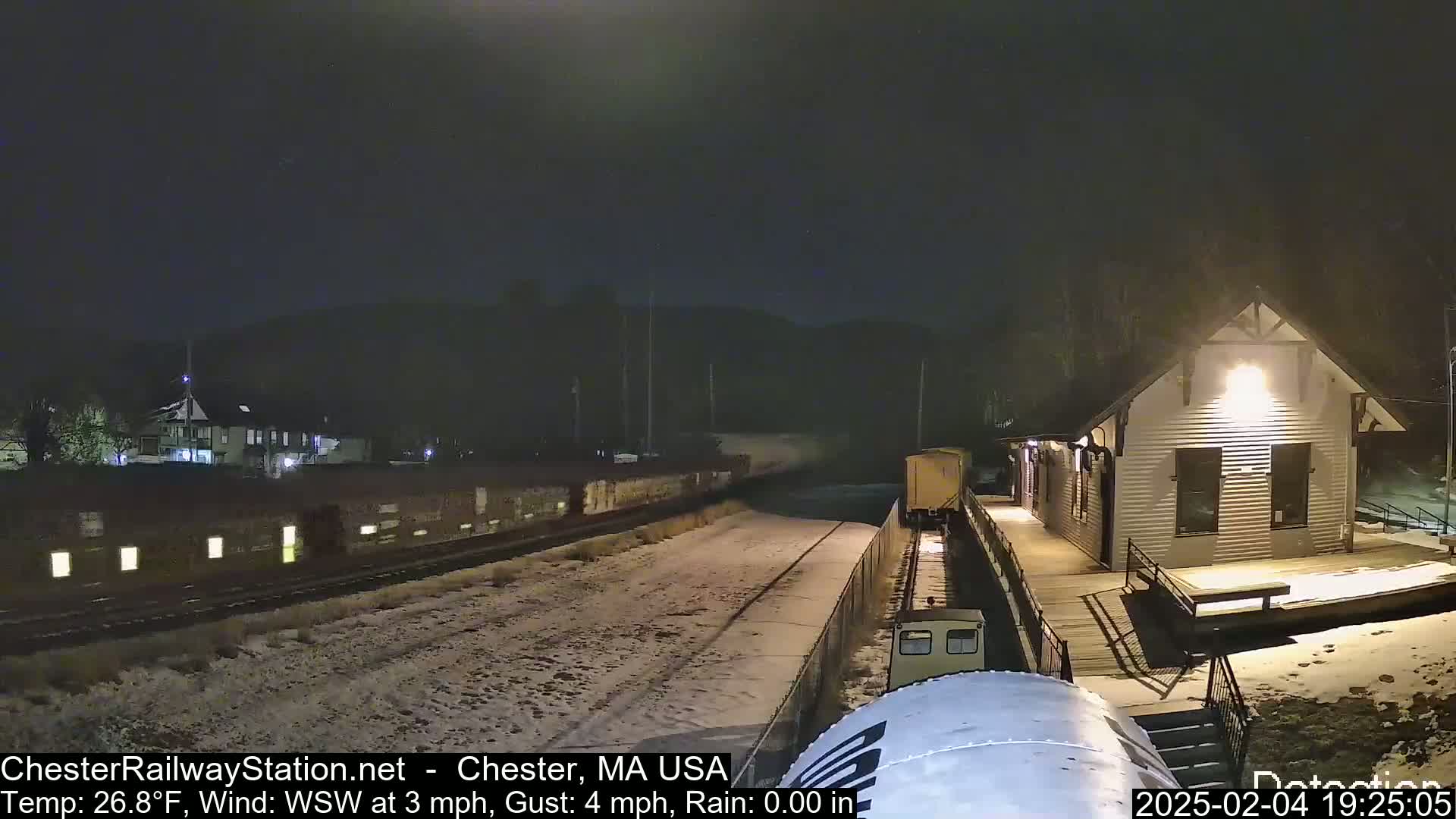 A snow-covered train track at night with a small train approaching a brightly lit train station.