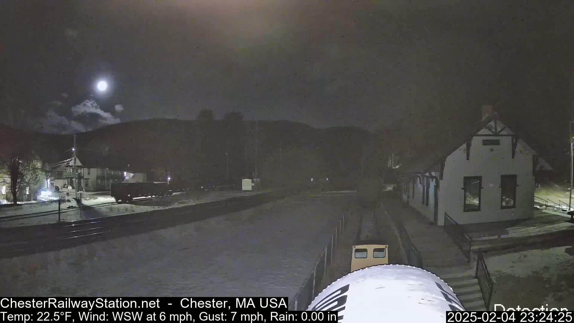A nighttime view of a train station and railway tracks, partially covered in snow, under a partly cloudy sky with a bright moon.