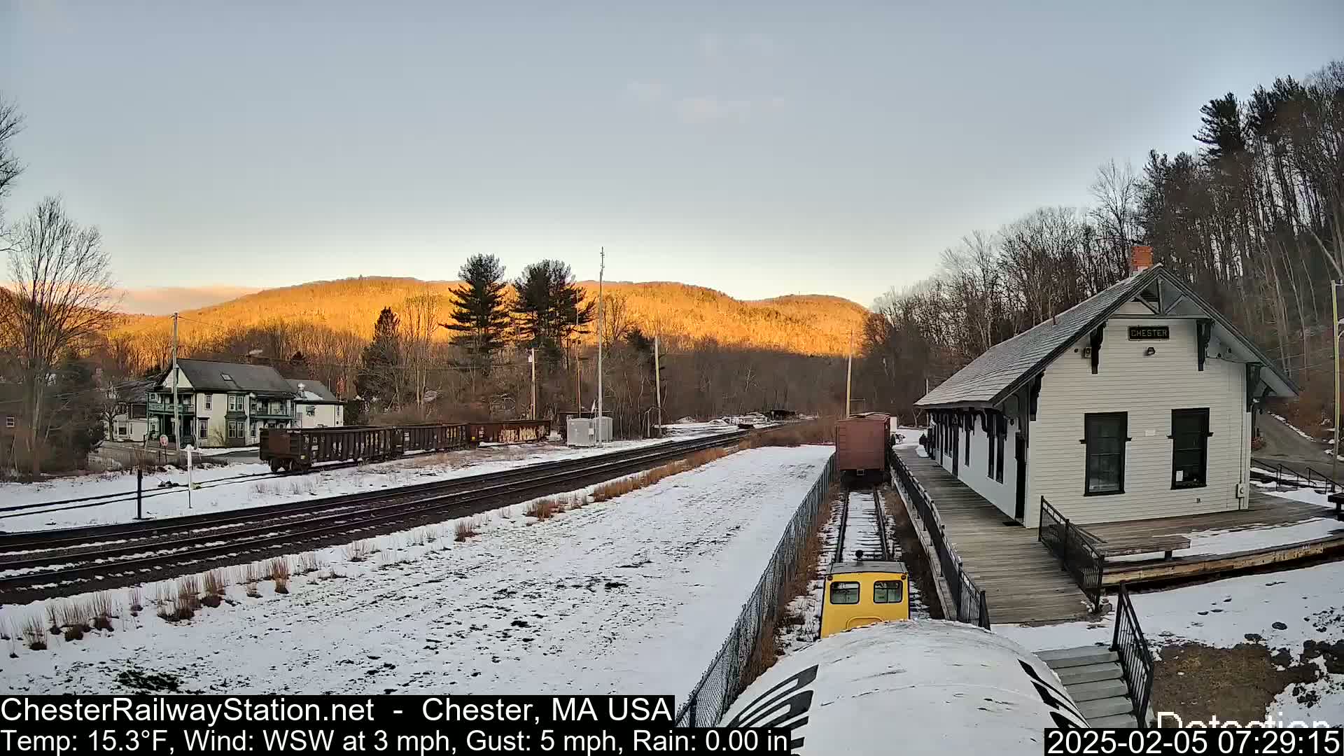 A snow-covered train station and tracks are seen at sunrise, with hills in the background illuminated by the rising sun.
