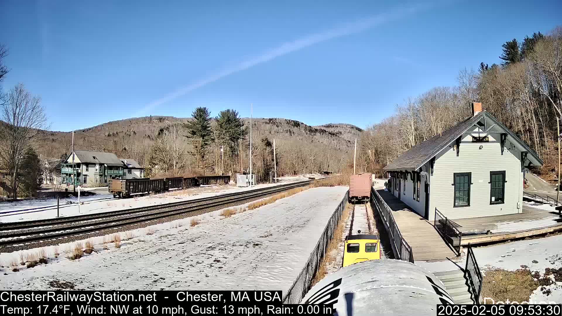 A train station and rail yard are visible on a sunny day with light snow cover, nestled between wooded hills.