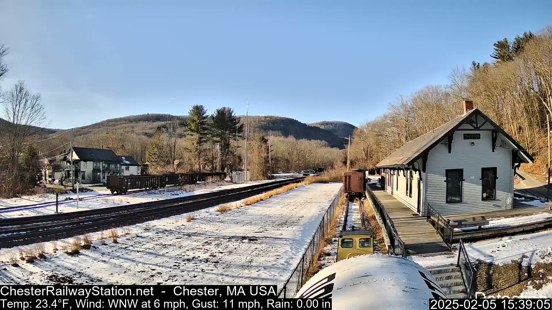 A snow-covered train station with a small train car sits beside a building, nestled in a valley between wooded hills under a clear, sunny sky.