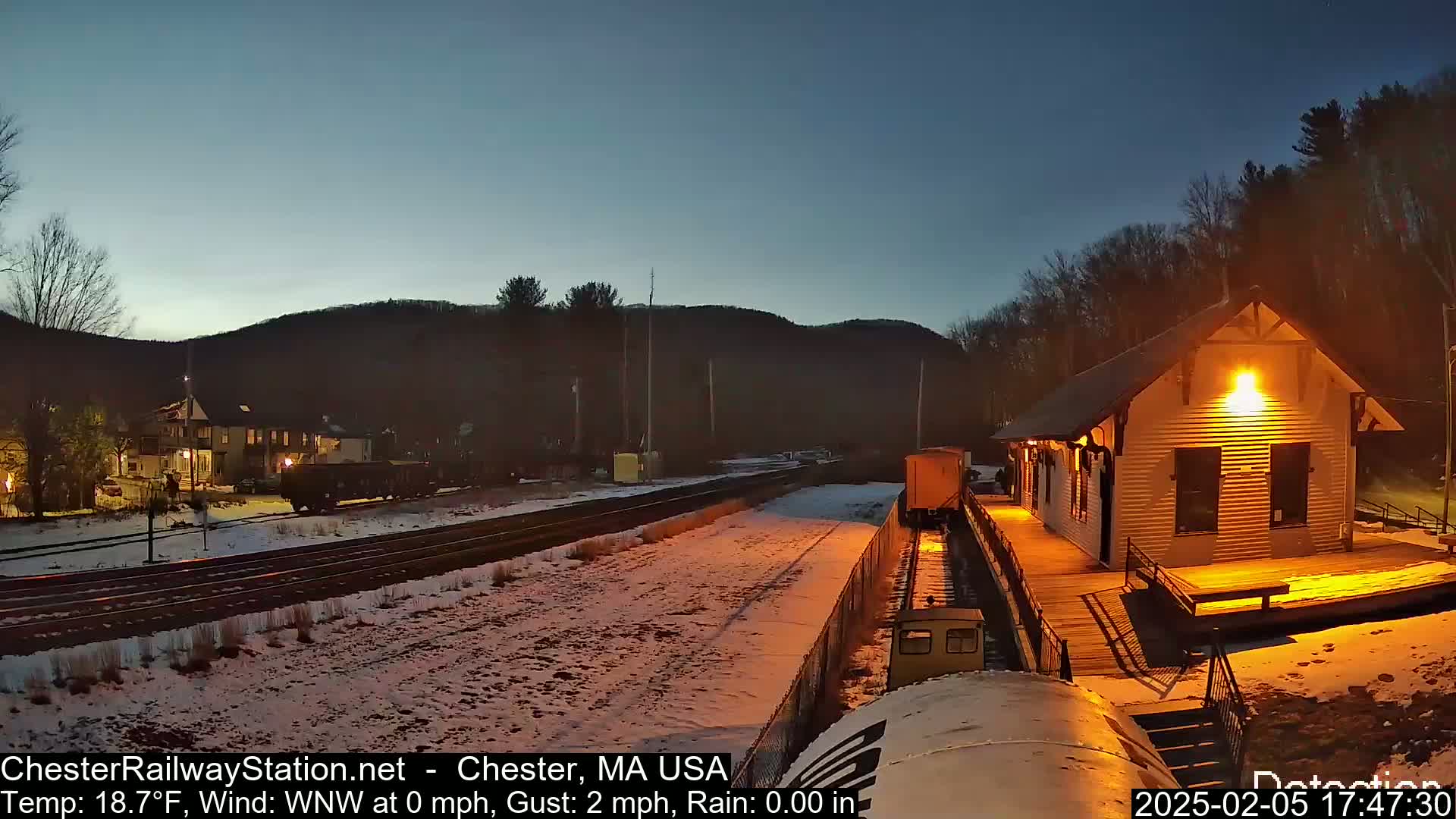 A snow-covered train station and rail yard are illuminated at twilight under a clear, dark blue sky.