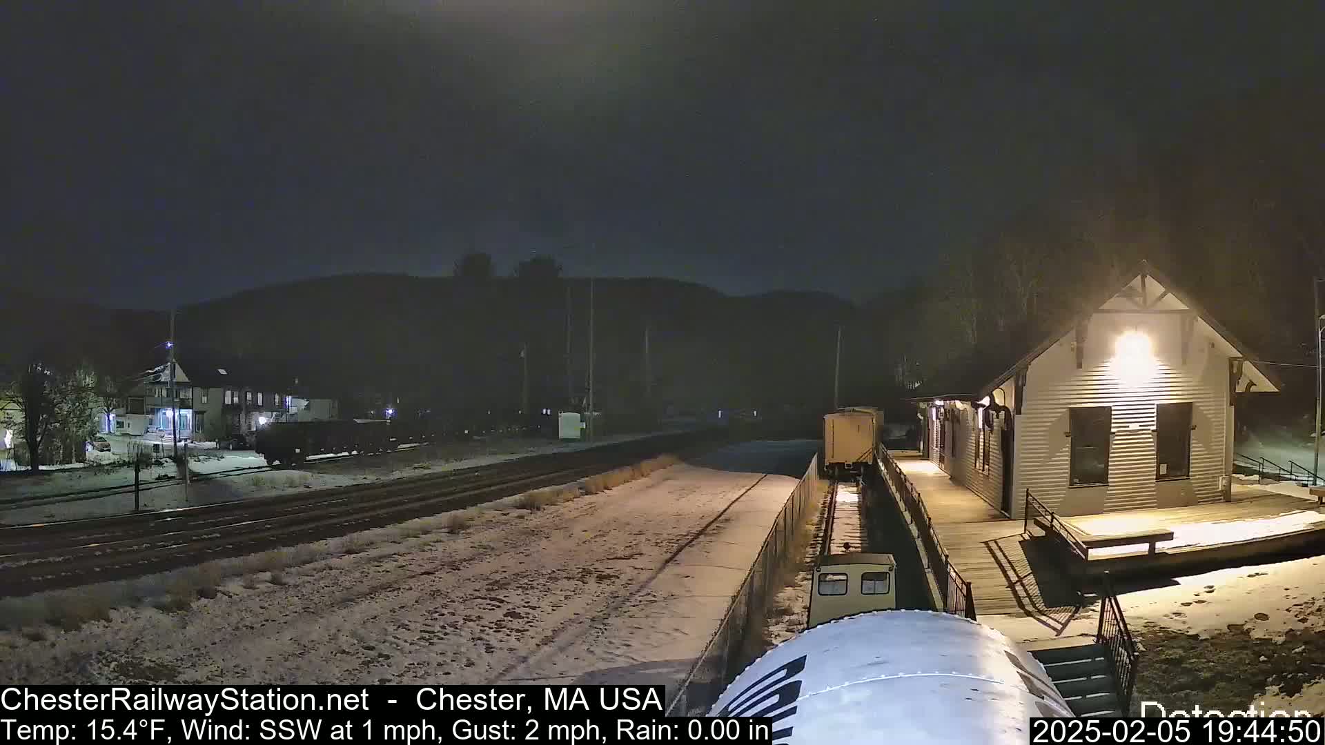 A train passes by a snow-covered railway station at night in light rain.