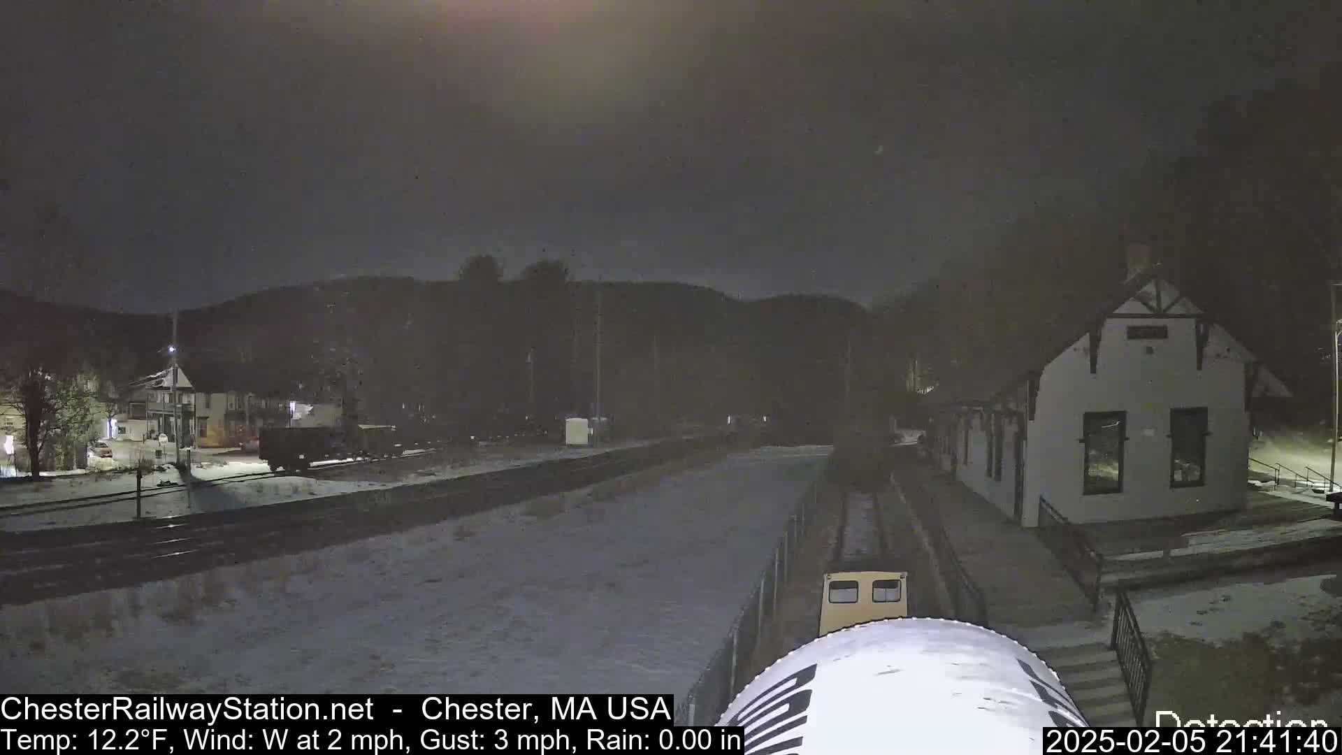 A nighttime scene of a train station and railway tracks lightly covered in snow under a dark, slightly snowy sky.