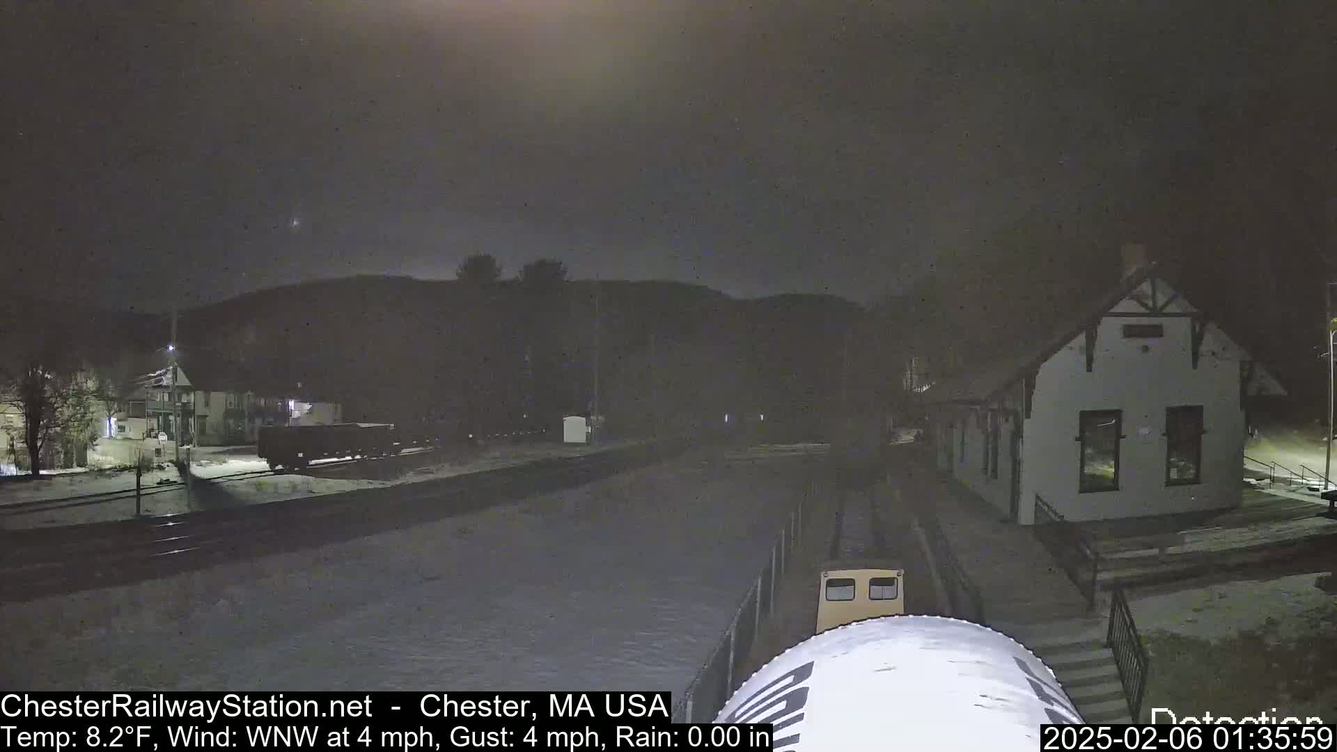 A nighttime view of a train station and rail yard with light snow cover under a dark, overcast sky.