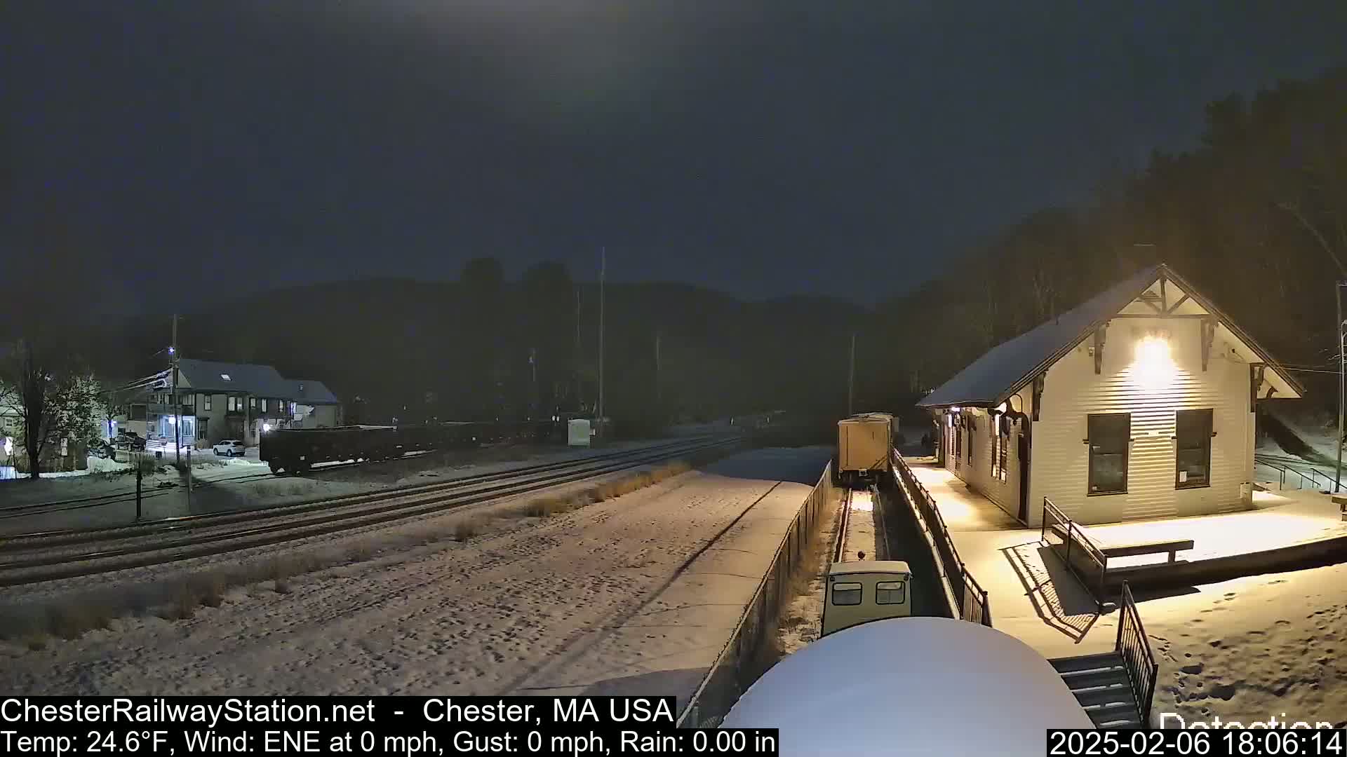 A snow-covered train station is illuminated at night, with a train partially visible on the tracks in the foreground.