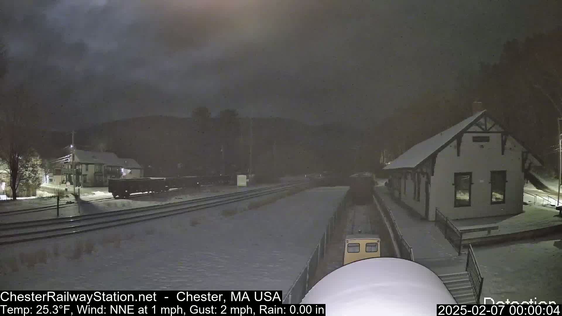 A train approaches a snow-covered railway station at night in light rain.