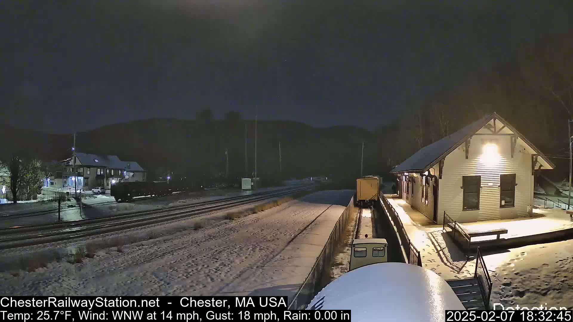 A snow-covered train station at night with a train partially visible on the tracks, under a dark sky with a WNW wind at 14 mph and gusts up to 18 mph.
