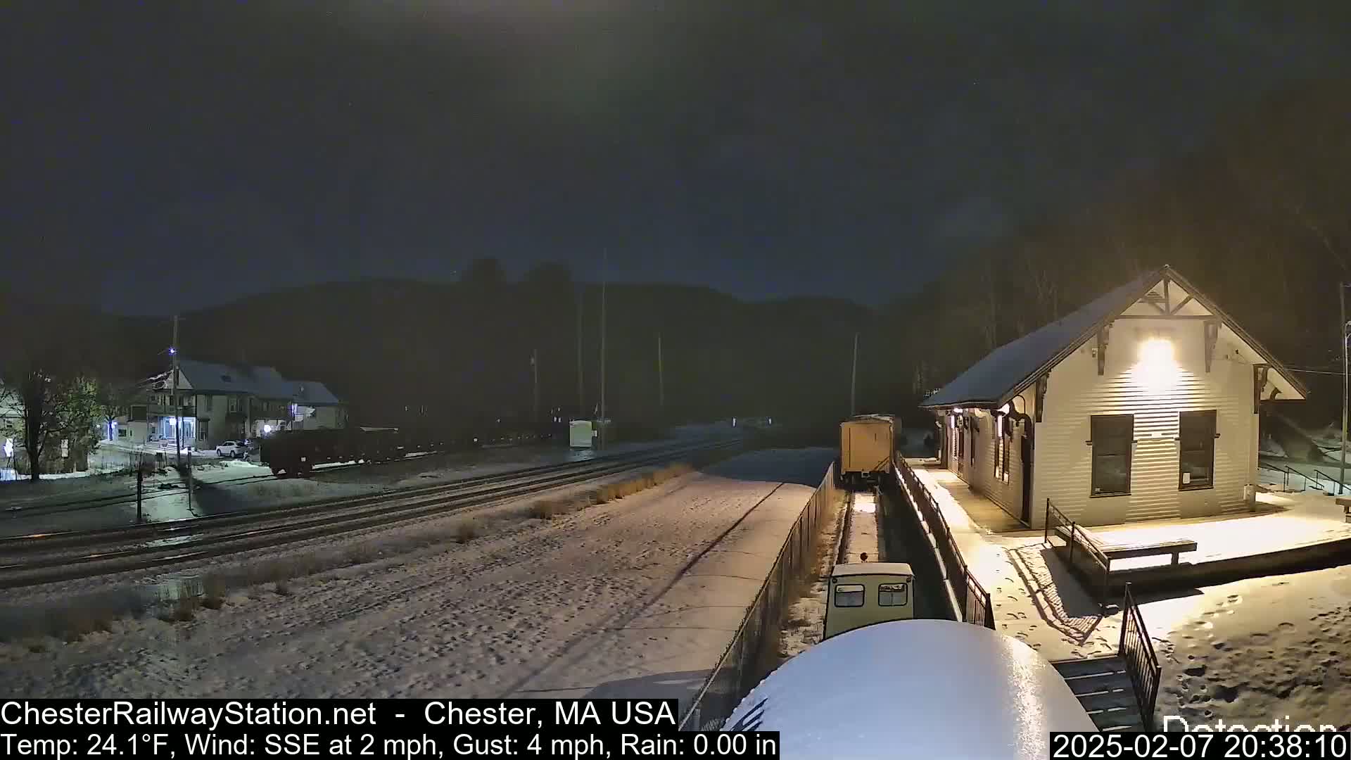 A snow-covered train station and railway tracks are visible at night under light rain.