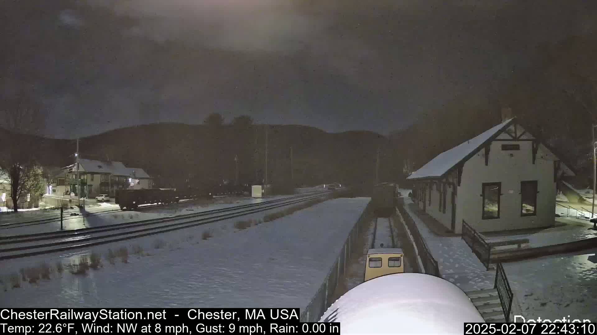 A snow-covered train station and rail yard are seen at night under a dark sky, with a small train approaching the station.