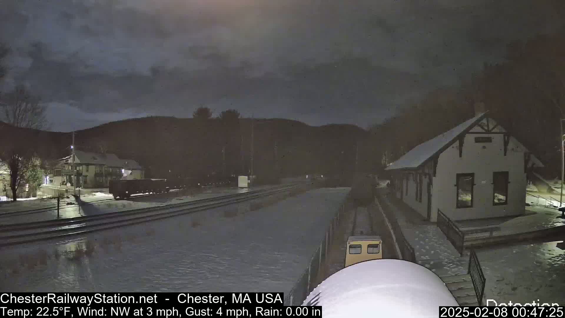 A nighttime view of a train station, railway tracks, and a small train car under a dark, cloudy sky with light rain and snow on the ground.