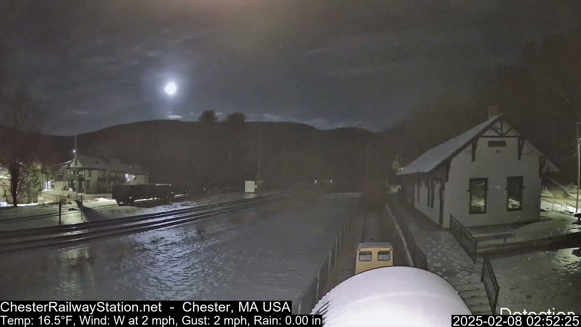 A nighttime view of a flooded railroad track near a small building under a bright moon and partly cloudy sky.