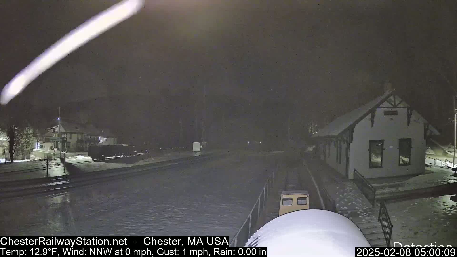 A nighttime view of a train station with light snow on the ground and a train car in the distance under a dark, slightly hazy sky.