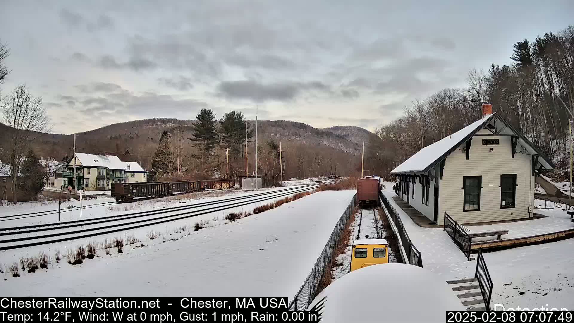 A snow-covered train station and tracks are seen on a partly cloudy day, with a small train approaching the station.