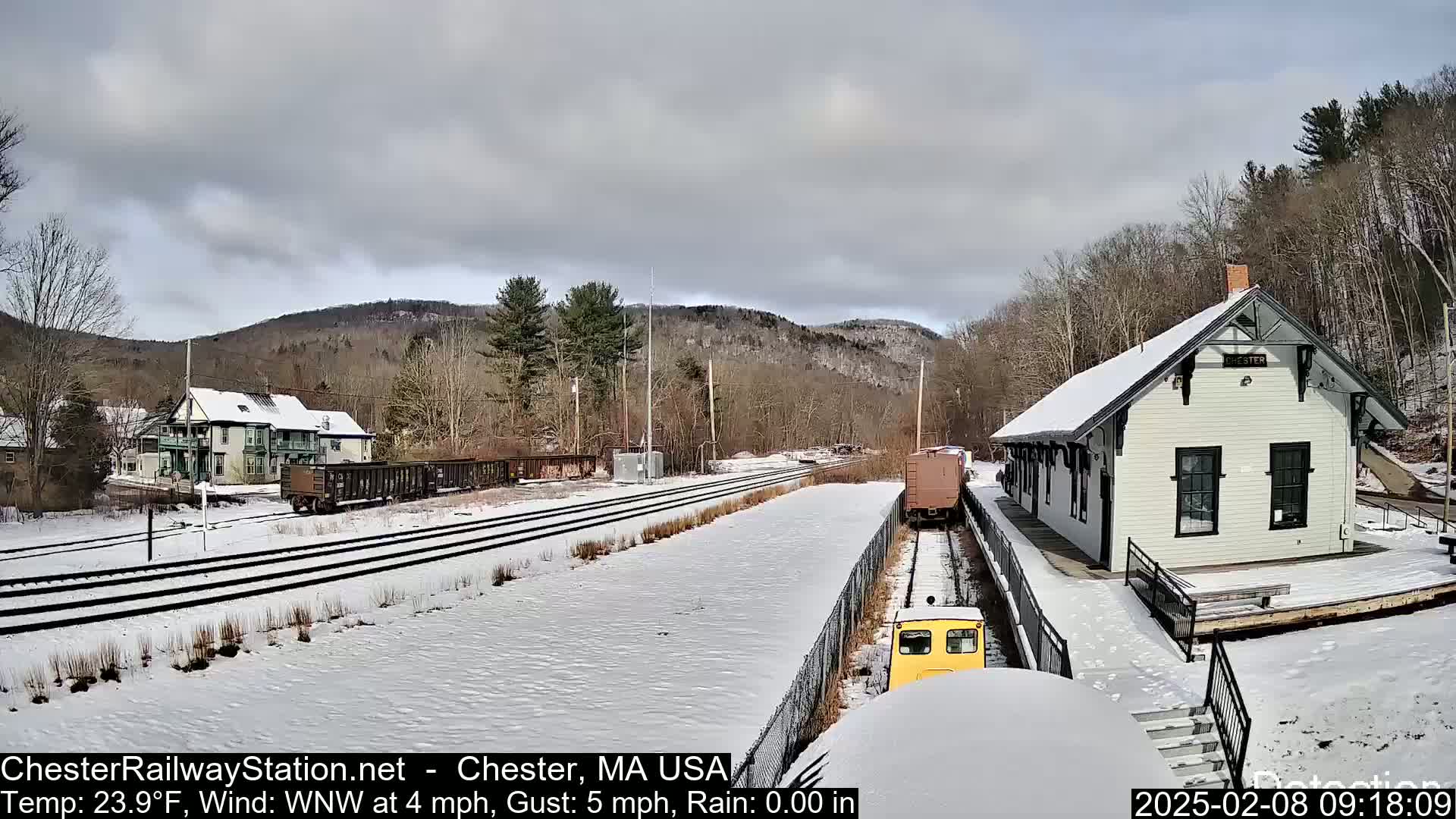 A snow-covered train station and railway tracks are visible, with a small yellow train approaching the station under partly cloudy skies.