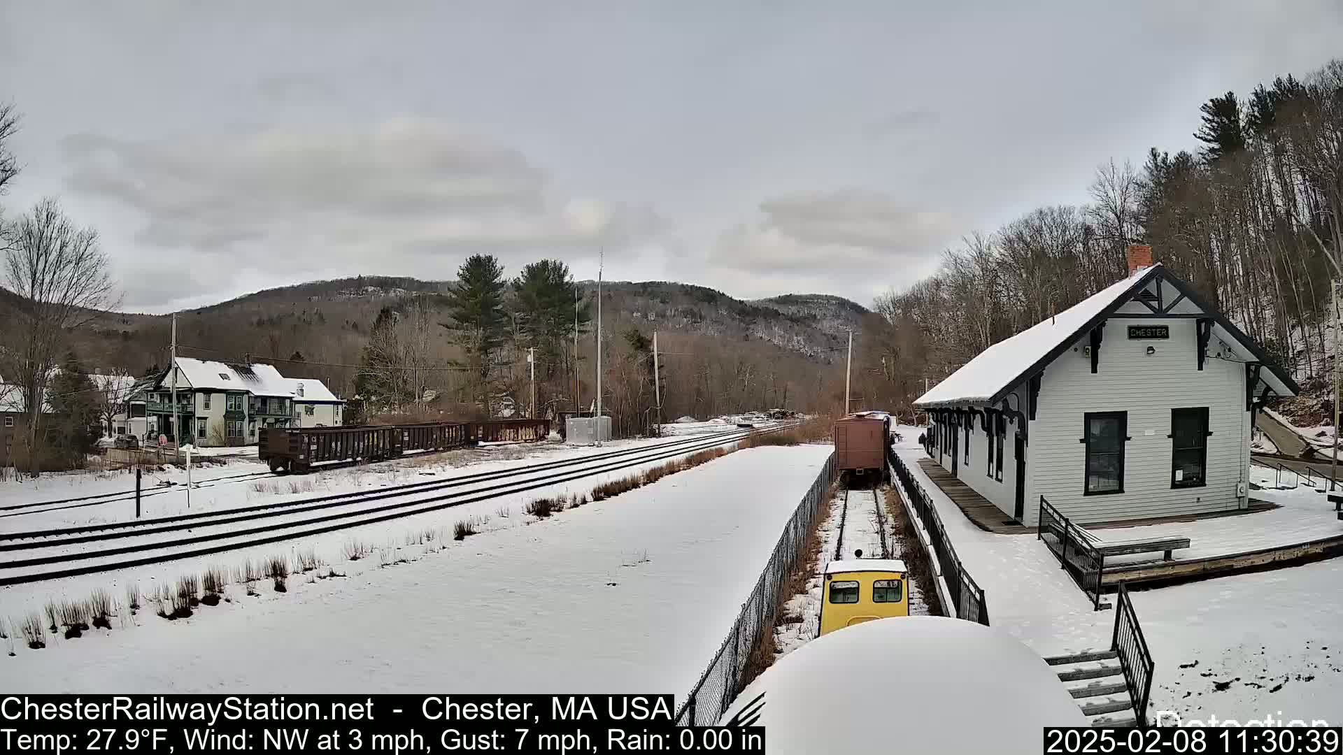 A snow-covered train station and railroad tracks are visible, with a train approaching the station under a cloudy sky.