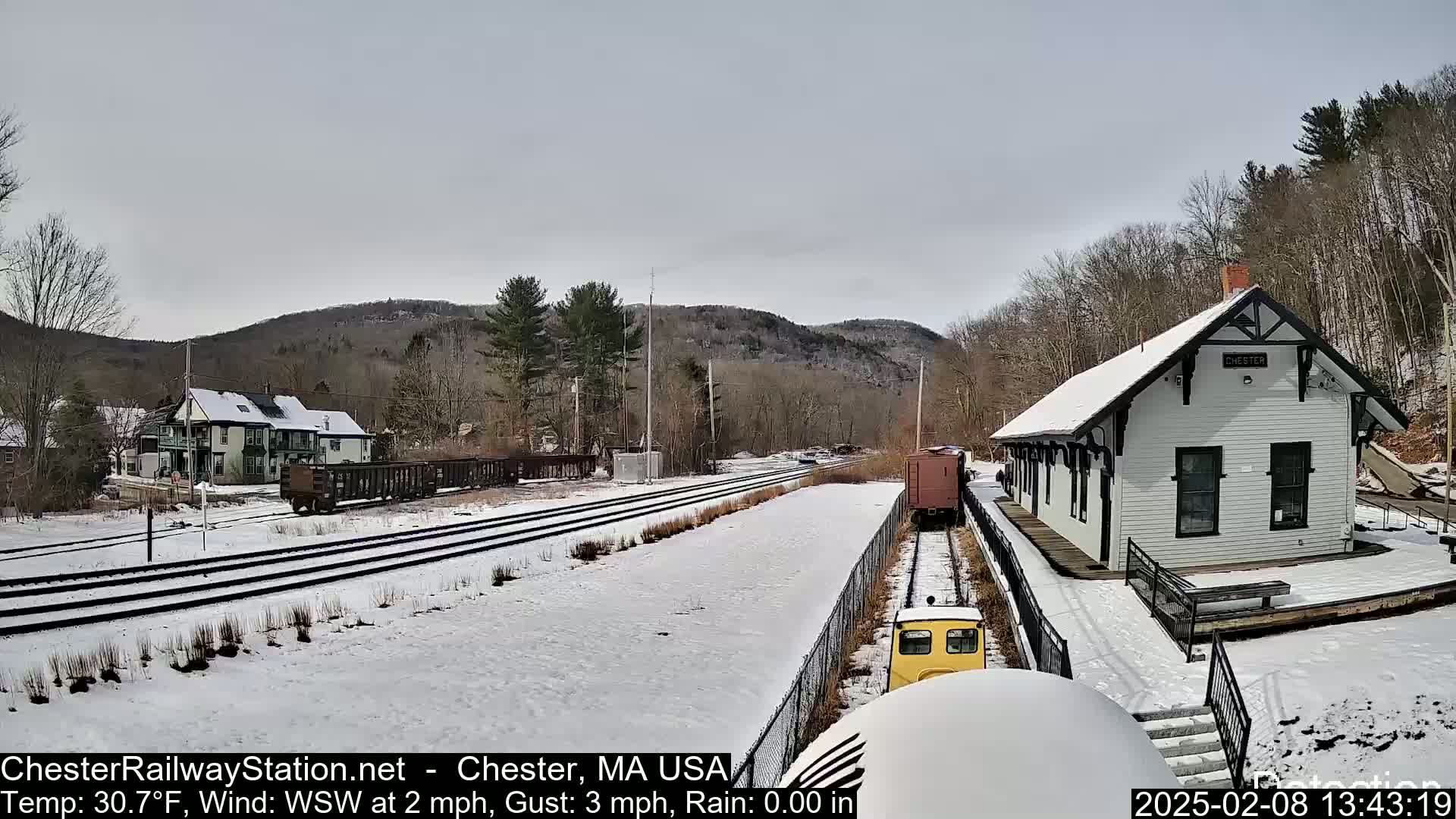 A snow-covered train station and railroad tracks are visible, with a small yellow train in the foreground and hills in the background under a cloudy sky.