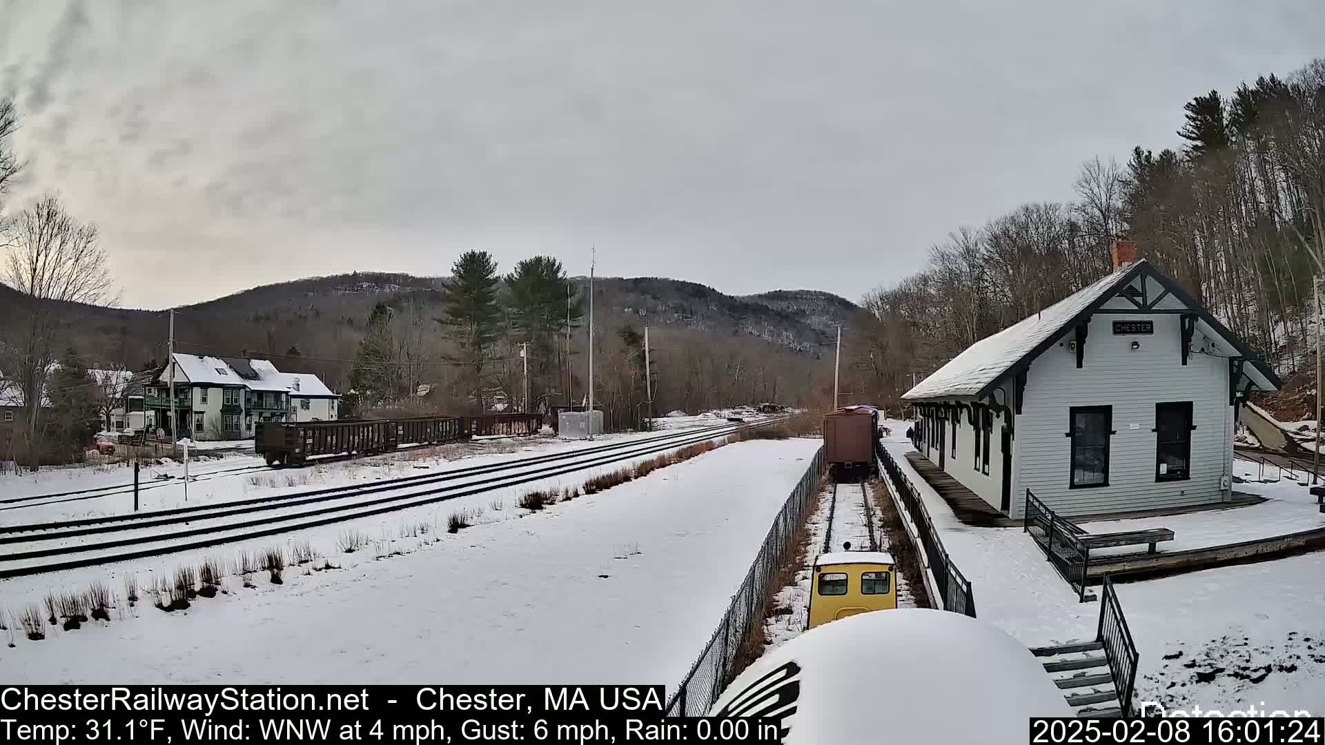 A snow-covered railway station and tracks are visible, with a small train car and freight cars nearby, under an overcast sky.