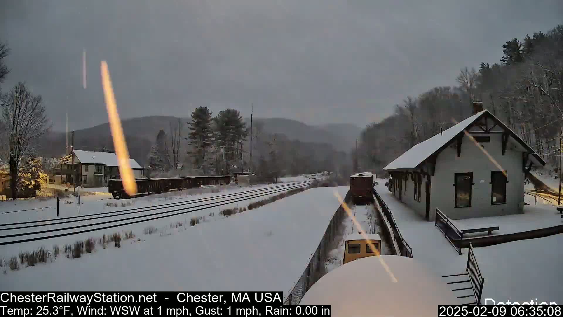 A snow-covered train station and rail yard are seen under an overcast sky, with a train partially visible in the foreground.