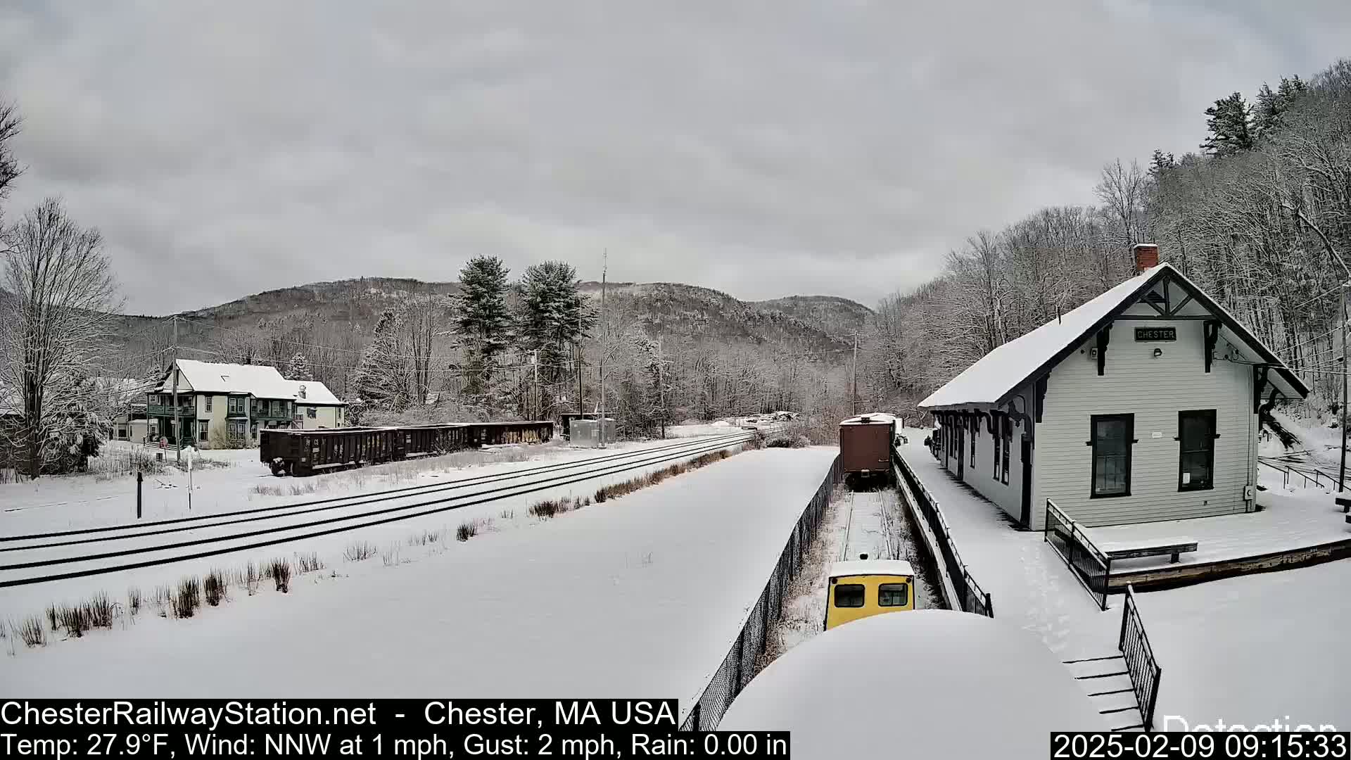 A snow-covered train station and rail yard are situated amongst snow-covered trees and hills under an overcast sky.
