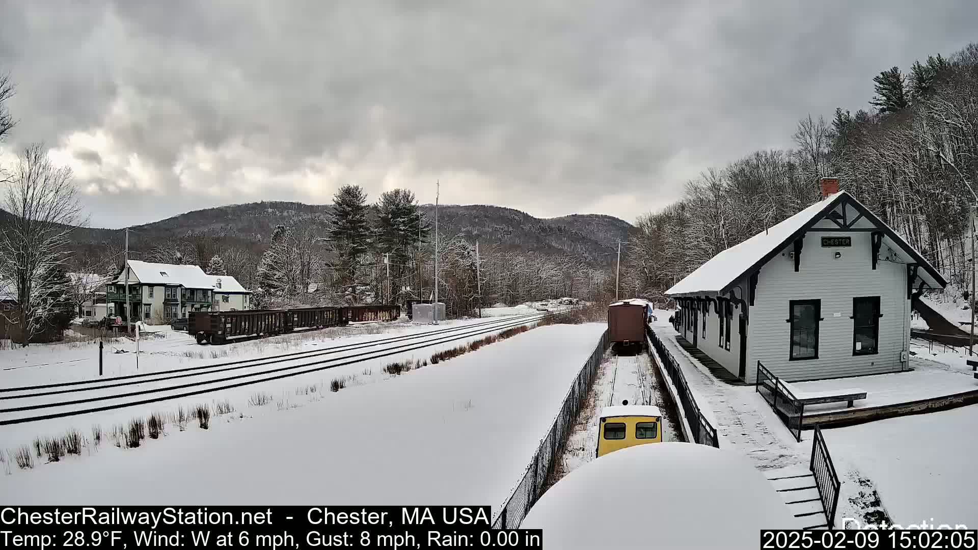 A snow-covered train station and railway line are visible under an overcast sky, with a train partially obscured by snow.